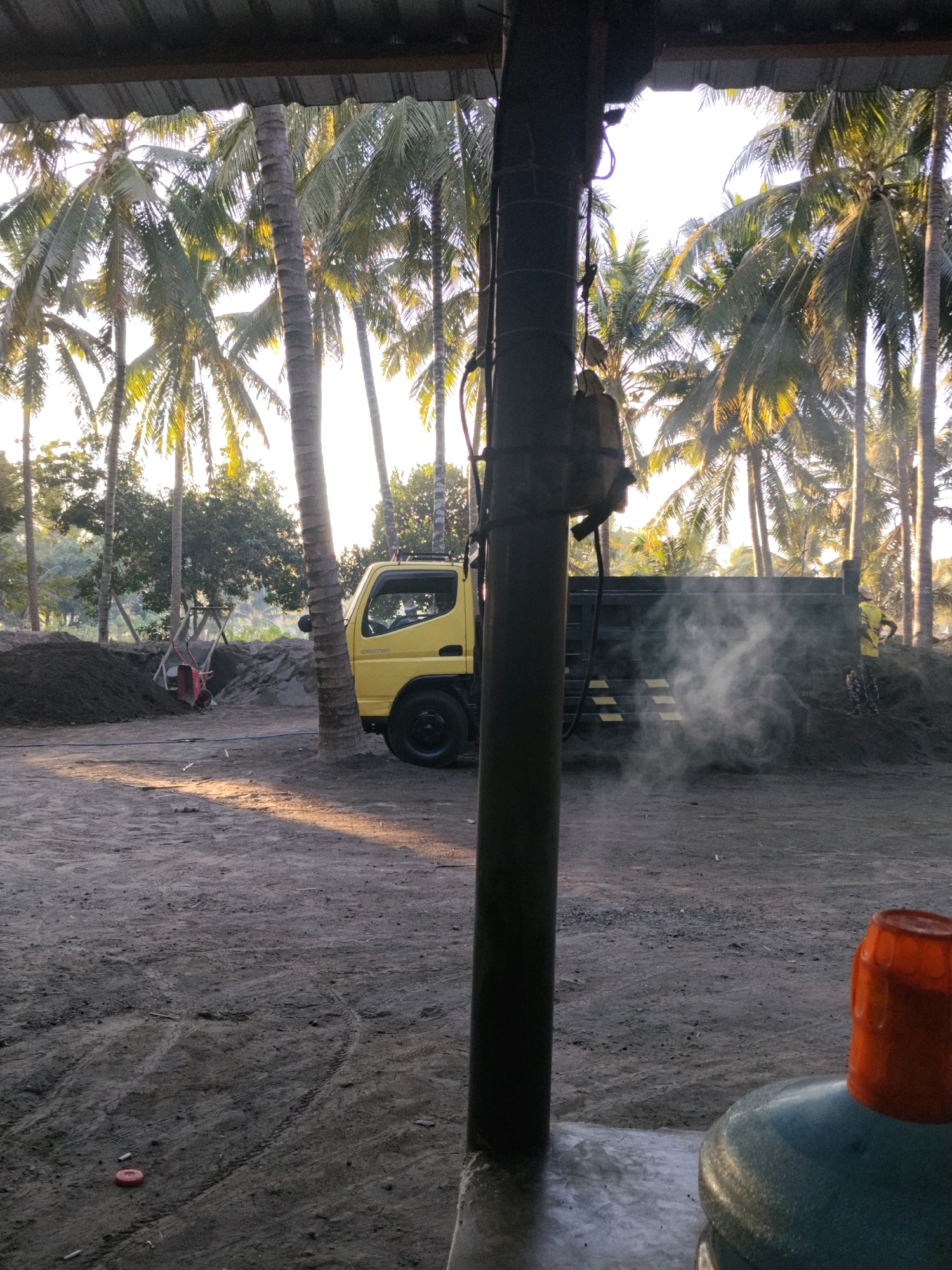This image depicts a scene in Ijobalit, Indonesia, likely at a construction or material processing site. In the foreground, a large dark pillar partially obstructs the view. Beyond it, a yellow dump truck is parked. The truck is emitting a significant amount of white smoke from its rear, obscuring part of the truck bed and the area behind it. A person wearing a yellow shirt and patterned pants can be seen standing near the rear of the truck, partially visible in the smoke.
The environment is characterized by a dusty, unpaved ground surface with visible tire tracks. Several large piles of dark material, possibly soil or aggregate, are present in the midground. To the left of the truck, a red wheelbarrow rests against a support structure. A grove of tall coconut palm trees dominates the background, with some smaller trees and foliage interspersed. The lighting suggests it is either early morning or late afternoon, with sunlight filtering through the palm fronds, casting long shadows on the ground. In the lower right corner, a large plastic water jug with an orange cap is visible. The overall impression is a busy, functional outdoor workspace.