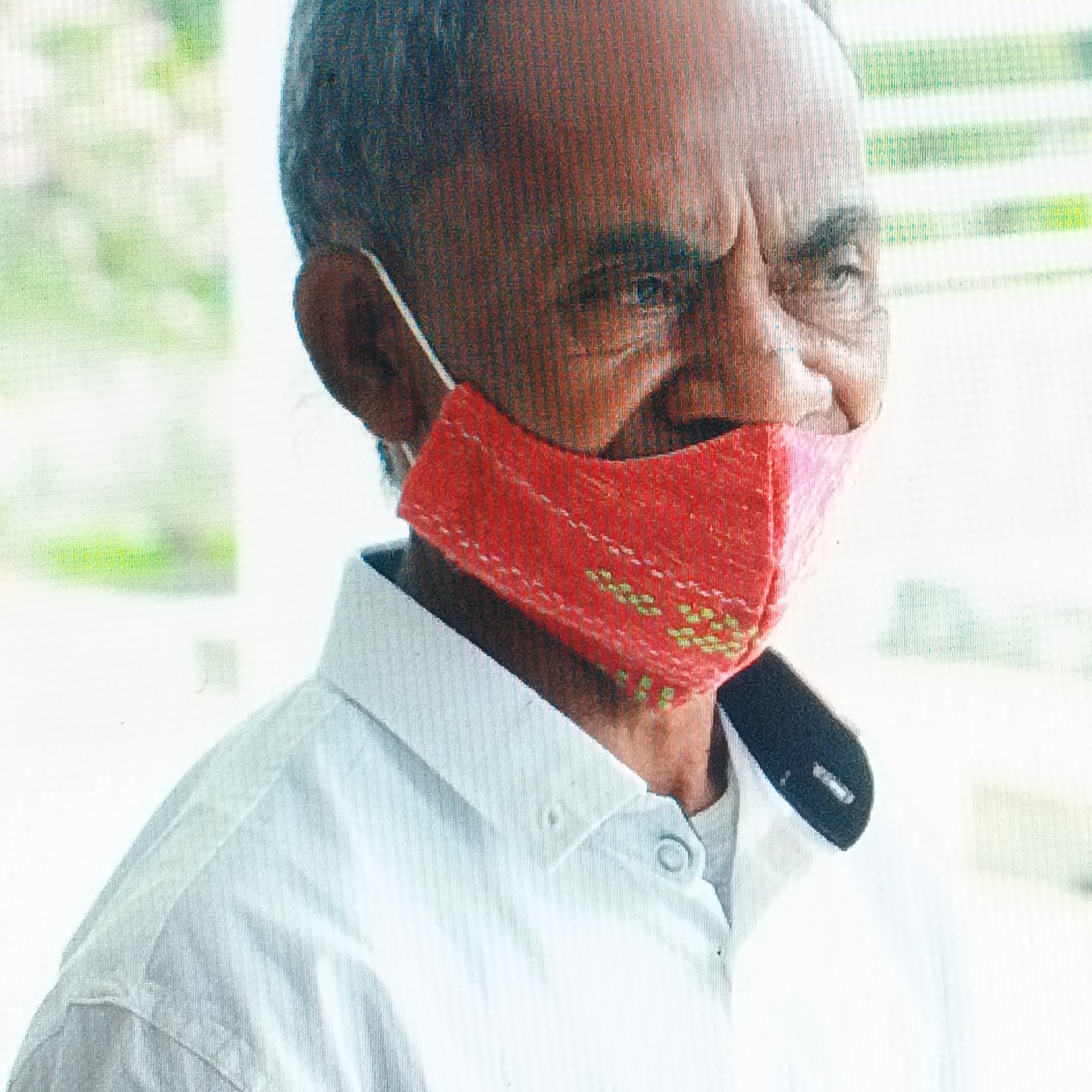 A headshot shows an older man from the chest up, looking to the viewer's left. He is wearing a white collared shirt, possibly a polo, with a red fabric face mask over his nose and mouth. The mask is embroidered with small, light green dots in a pattern. The man has dark hair, visible on the sides of his head and thinning at the top, and appears to have weathered skin. He has dark eyebrows and his eyes are visible above the mask. The background is blurred with green and white horizontal lines, suggesting an outdoor setting or a room with blinds. There is no identifiable information in the image indicating the specific location in Ende, Indonesia.