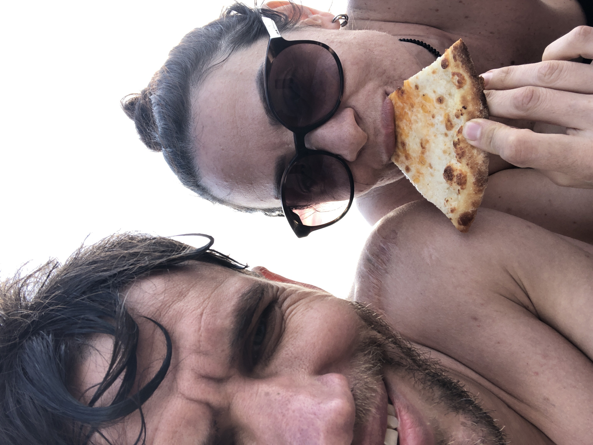A close-up photograph depicts two individuals, a man and a woman, positioned in a tight frame, likely a selfie. The background is completely overexposed white, providing no discernible environmental details or specific geographical context to confirm the location as Sestroretsk, Russia.
The man, positioned on the left and slightly to the foreground, presents his left profile from the mid-forehead down to his upper chest. He has dark, wet hair, a short beard, and is smiling broadly, revealing white teeth. His left eye is visible, directed towards the camera. His skin appears tanned, and faint red marks are visible on his left shoulder and upper arm.
The woman, positioned on the right, faces towards the camera, with her face, right shoulder, and right hand visible. She has dark hair styled in a small bun on top of her head. She is wearing large, dark-lensed sunglasses that obscure her eyes. A silver-colored hoop earring is visible in her left ear, and a thin, dark necklace is around her neck. Her skin also appears tanned. She is actively biting into a triangular slice of cheese pizza, which she holds with her right hand. The pizza slice has a slightly browned crust. Both individuals appear to be outdoors or in a brightly lit setting, possibly in minimal clothing.