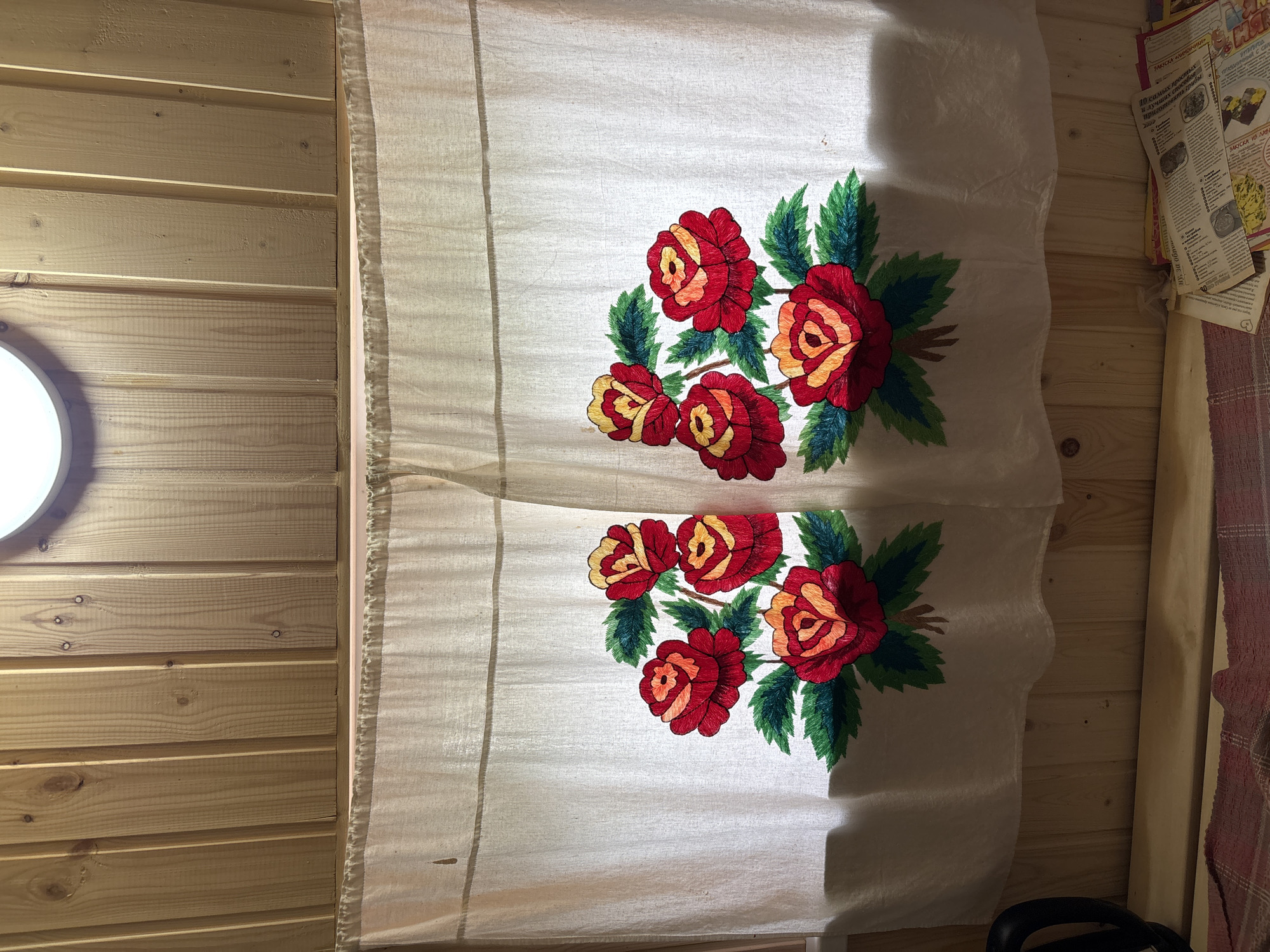 The image shows a close-up of a white curtain with embroidered red and yellow roses and green leaves. The curtain is hung in front of a wooden wall. A light fixture is visible at the top center of the image, casting a warm glow. In the lower part of the image, a wooden shelf is visible, with some newspapers and a red and white checkered cloth on it. The scene appears to be in a rustic indoor setting. There are no people visible in the image. The city of Ильпанур, Russia is not depicted in this image.