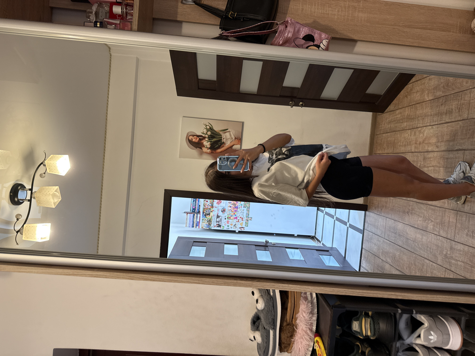 A young woman with long dark hair is standing in a room and taking a mirror selfie. She is wearing a white t-shirt and black shorts. She is holding a phone in her right hand. In the background, there is a door, a shoe rack with shoes, and a wall with a picture of a woman holding flowers. There are also shelves with perfumes and bags. The room appears to be a bedroom or a dressing room. The floor is made of wooden planks. The lighting in the room comes from a ceiling fixture with multiple lights.