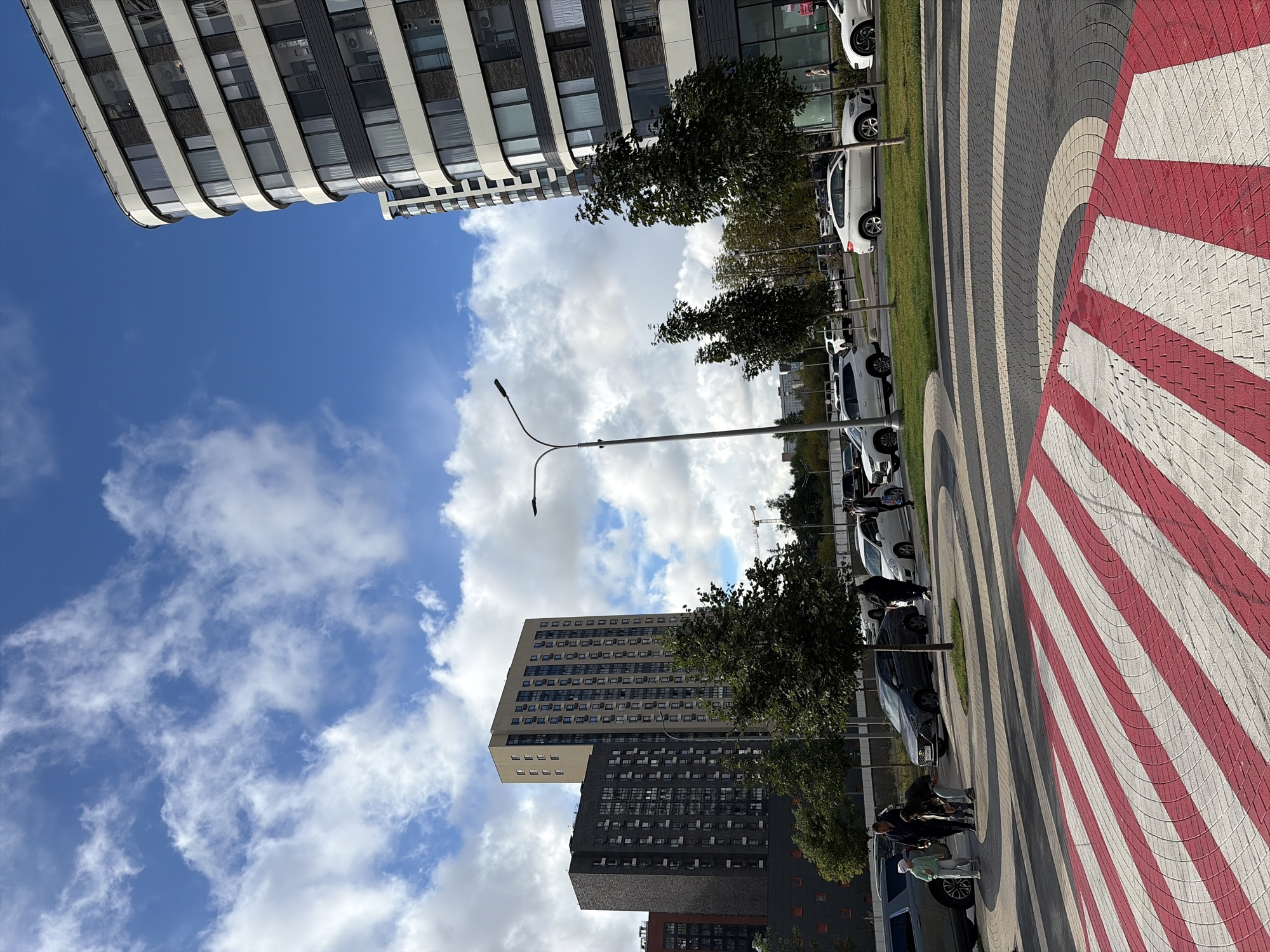 This image depicts a modern urban scene with multiple residential buildings and parked cars under a cloudy blue sky. The perspective is from a low angle, emphasizing the foreground pedestrian crossing with red and white stripes.
In the foreground, a distinct pedestrian crossing is paved with red and white stripes. Beyond the crossing, a curved street is visible with several cars parked along the side. Among the parked vehicles are a dark grey SUV, a dark grey sedan, and several white cars including what appears to be a white BMW X-series.
Several people are present. A group of four individuals, including two adults and two children, are standing near the dark grey SUV. Further down the street, a solitary figure is walking away from the camera. Another person is visible near the entrance of one of the buildings.
The background features several tall residential buildings. To the right, a curved, multi-story building with a facade of white and dark grey panels and numerous windows dominates the skyline. To the left and center, two rectangular residential towers are visible. One is a lighter beige color and taller, while the other is darker grey and appears slightly shorter. Trees are strategically planted along the street, providing some greenery. A streetlamp is also present in the mid-ground.
The sky is blue with scattered white clouds. The overall environment suggests a well-developed residential area within a city. Based on the architectural style of the buildings, particularly the curved facade of the building on the right, and the presence of street signs and infrastructure consistent with Russian urban planning, this scene is likely located in Moscow, Russia. Specifically, the modern, large-scale residential complexes are characteristic of newer developments in Moscow.