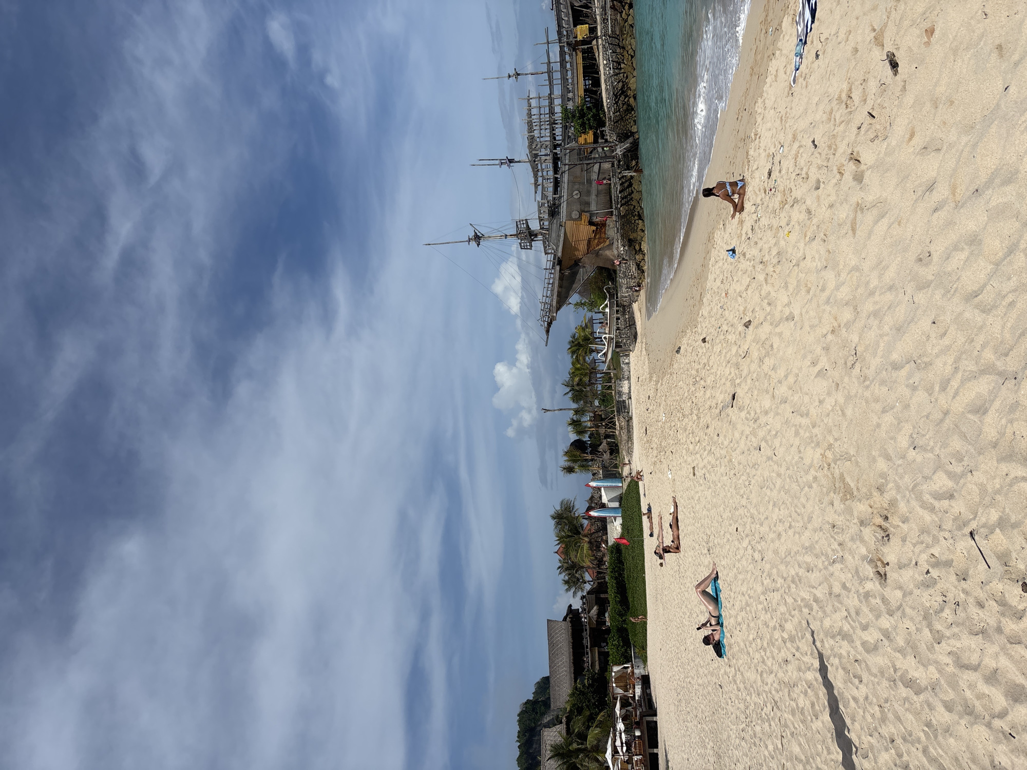 The image shows a beach scene in Kuta Selatan, Indonesia. A replica pirate ship is anchored near the shore. The beach is sandy, with people sunbathing and wading in the water. The sky is blue with scattered clouds. Buildings and trees are visible inland.