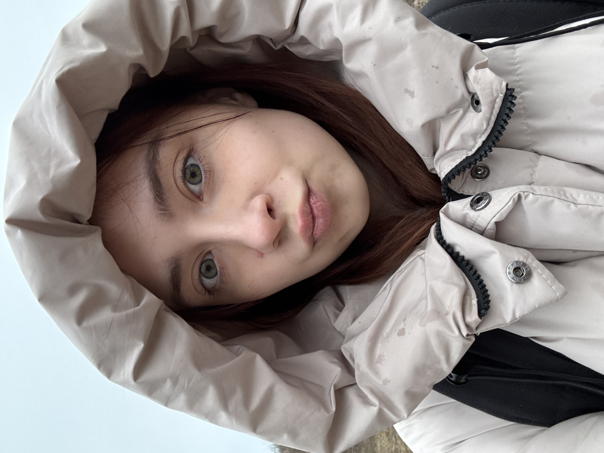 A close-up, slightly rotated, eye-level shot features a young woman in a beige hooded jacket. She has fair skin, reddish-brown hair, and green eyes. The hood is large and frames her face, with some splotches visible on the fabric. A black backpack strap is visible near her right shoulder. The background is blurred, possibly indicating an outdoor setting with a hint of sky. The location is Chaikovsky, Russia.