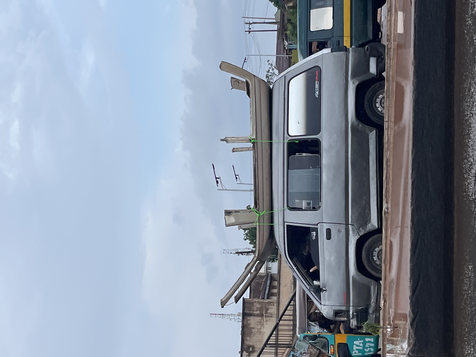 A gray van is driving on a street with a boat strapped to its roof. A person is visible in the driver's seat of the van, and another person is visible in the driver's seat of a green van parked next to it. The scene is set in Gura Top, Nigeria, with buildings and utility poles in the background. The sky is overcast.