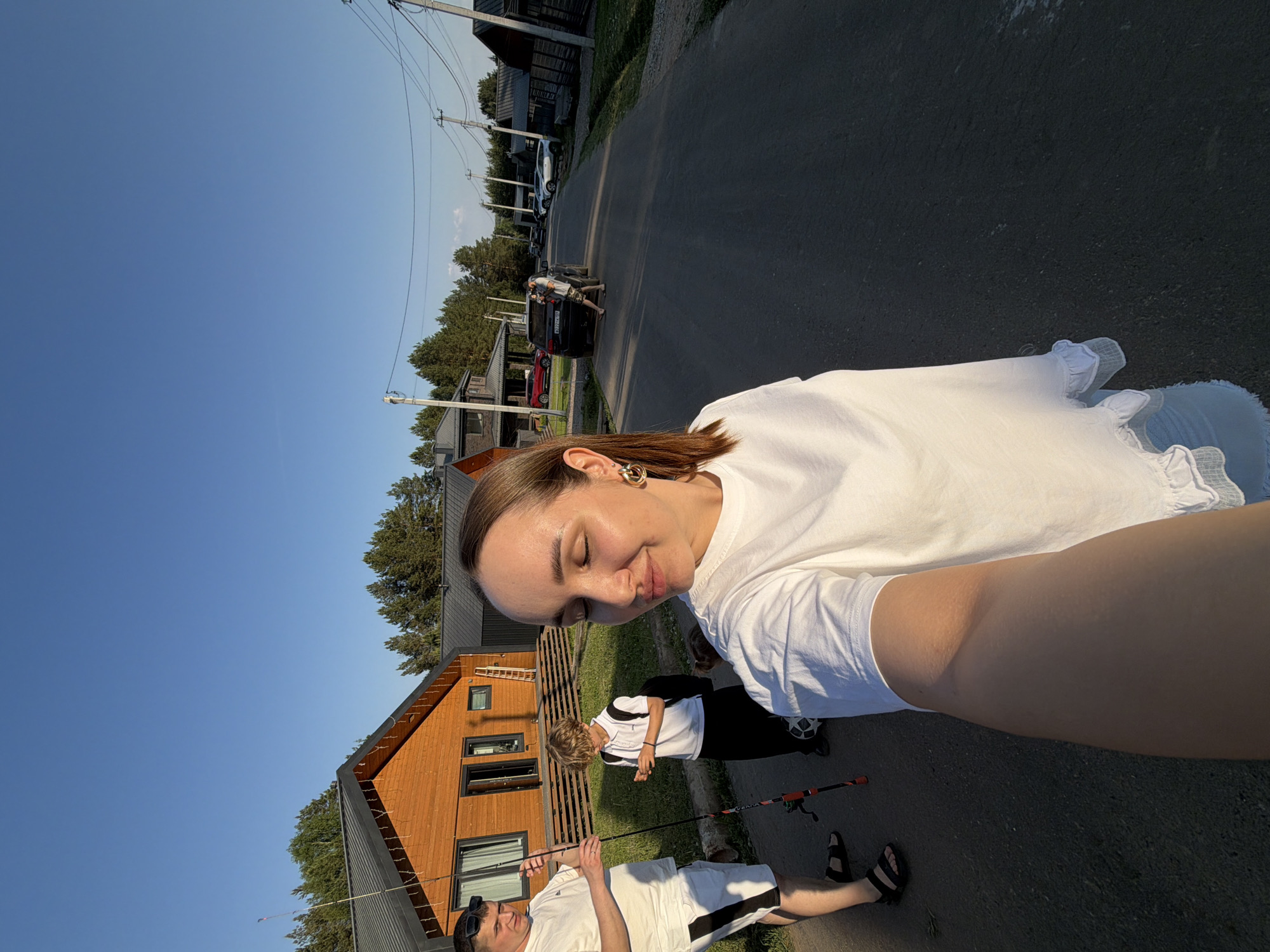 The image shows a selfie-style photograph taken outdoors. The primary subject is a woman with light skin, light brown hair pulled back from her face, and gold hoop earrings. She is wearing a white T-shirt. Her eyes are closed, and she has a slight smile.
In the background, two other individuals are present. One is a man wearing a white shirt and dark sunglasses, holding a fishing rod. The second person is a younger individual wearing a white shirt and black pants. A black and white soccer ball sits on the ground next to them.
Behind these figures is a tan building with dark accents and a ladder leaning against it. The setting appears to be in a residential area of Kamyennoye, Russia, with a paved road and other houses visible in the background. Cars are parked along the road. Utility poles and wires stretch across the top of the frame. The sky is clear and blue.