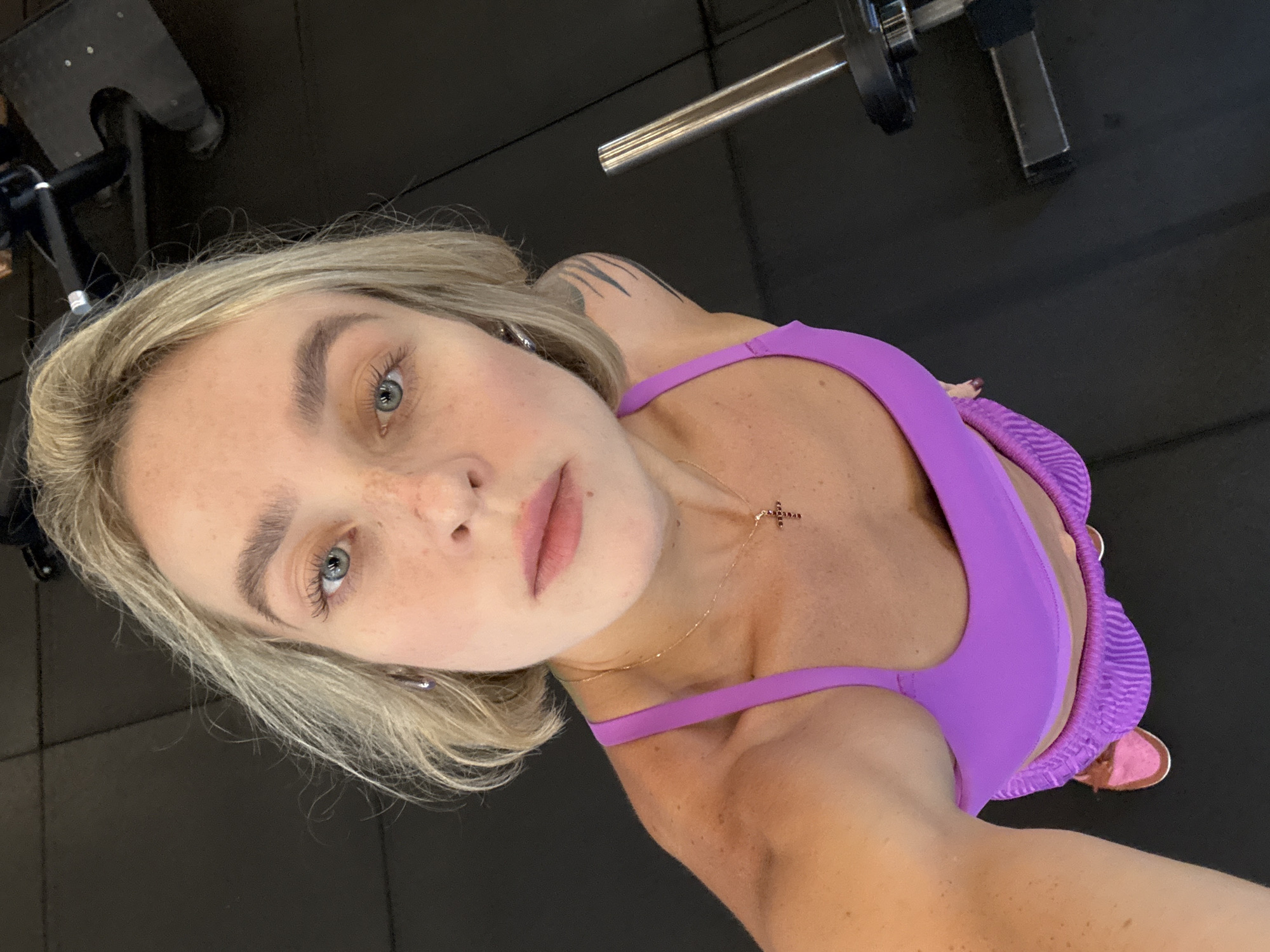 An overhead, medium shot captures a young Caucasian woman with blonde, chin-length hair and blue eyes in a gym. She is wearing a bright purple sports bra and matching striped shorts. A delicate gold necklace with a cross pendant rests on her collarbone. Her left arm is visible, showing a tribal tattoo on her upper arm and shoulder. The background is a dark gym floor, with a portion of a weight-lifting machine visible on the right side of the frame. The lighting is bright, highlighting the woman's features and the vibrant colors of her attire. The image is a selfie, taken from a high angle. The woman's expression is neutral, with her gaze directed towards the camera. The scene is devoid of any specific geographical markers that would indicate a location in Rawai, Thailand.