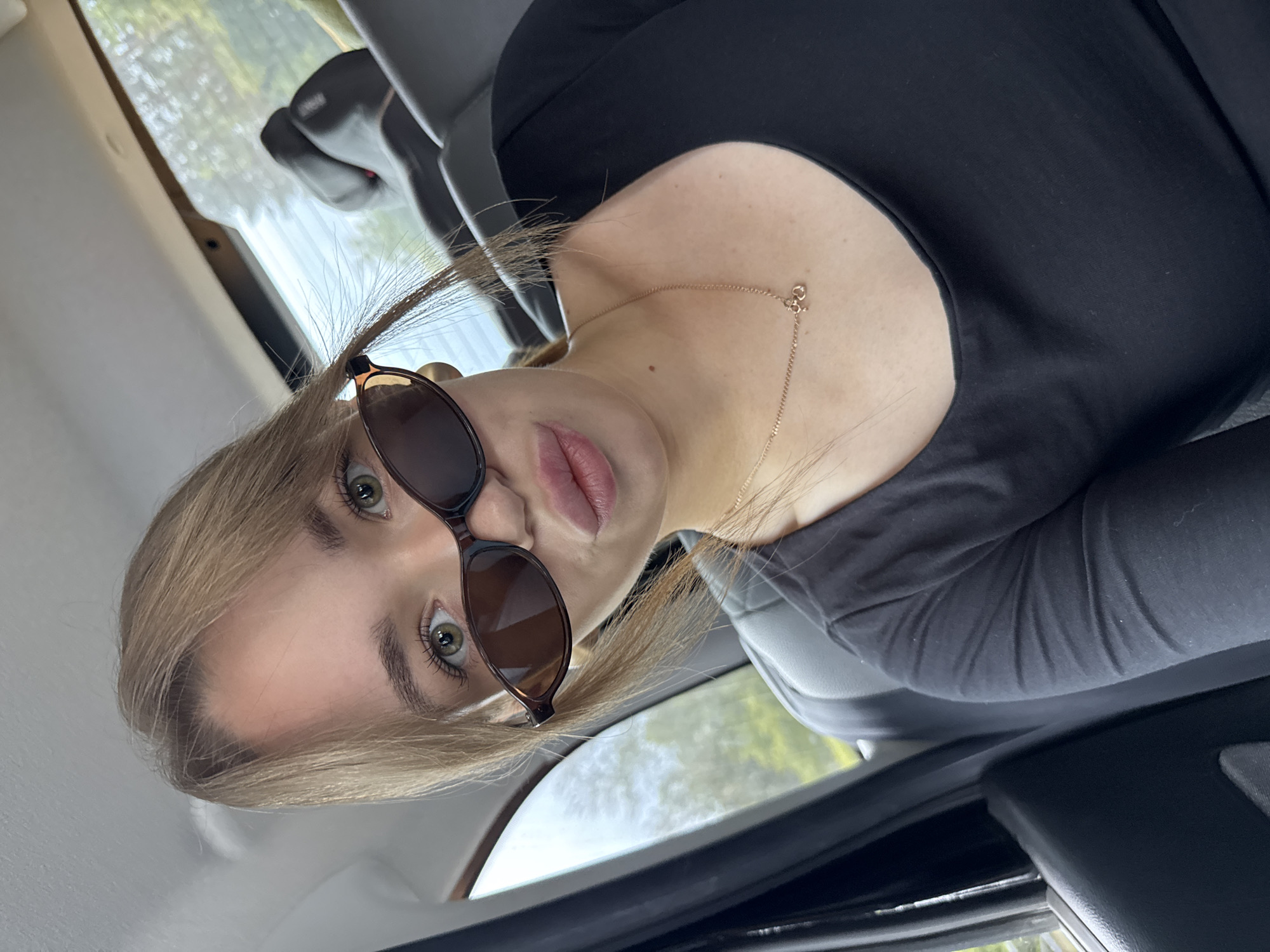 The image shows a young woman with blonde hair and green eyes, wearing sunglasses and a black shirt. She is inside a vehicle, likely a car, with the back seat visible behind her. The woman is looking at the camera with her lips slightly pursed. The background shows trees and foliage outside the car window, indicating an outdoor setting. There are no discernible landmarks that would specifically place this image in the city of Chaikovsky, Russia.