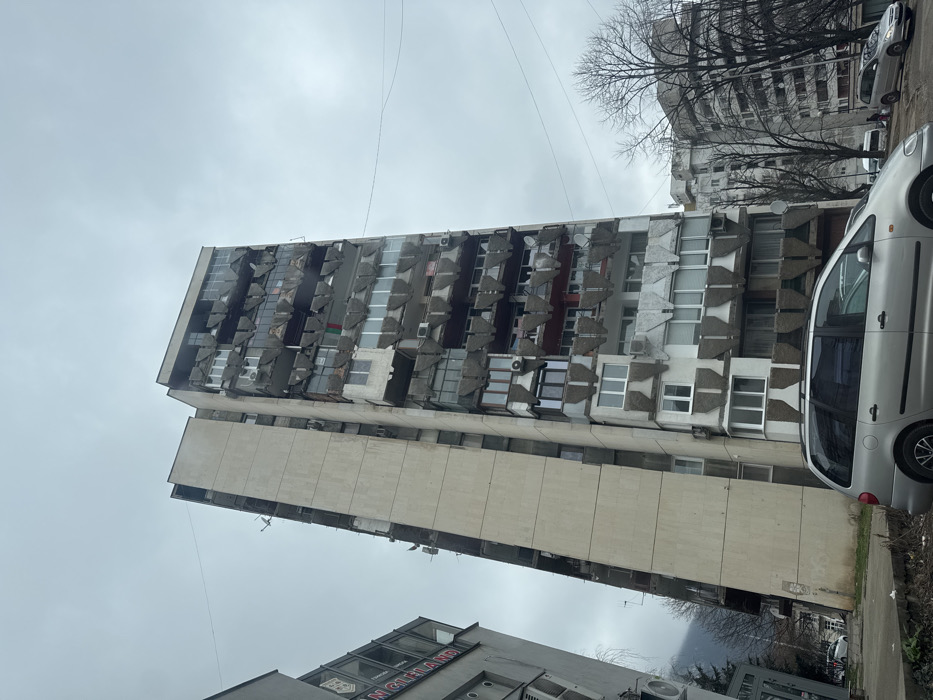The scene depicts a tall, multi-story concrete residential building located in Vratsa, Bulgaria, under an overcast sky. The building, exceeding twelve floors in visible height, features a flat, light beige/grey concrete side facade on the left. Its front facade is characterized by numerous balconies, many of which have distinctive, dark grey, fan-shaped concrete railings or screens. Numerous balconies show evidence of resident modifications, including enclosed sections with various window types, predominantly white PVC frames.
Exterior features on the building include multiple air conditioning units attached to walls and balconies, and several satellite dishes. A green, white, and red flag is draped from a balcony on one of the mid-upper floors. The concrete surfaces exhibit signs of weathering and minor discoloration.
To the left, a portion of a shorter, lighter-colored building is visible, featuring a dark-lettered sign with a circular logo, indicating a commercial establishment. In the distance to the right, another multi-story residential building, similar in architectural style, is partially obscured by bare deciduous trees.
In the lower foreground, a silver minivan, possibly a Volkswagen Sharan or similar model, is parked facing right, displaying roof rails. Other vehicles are visible parked along a street further in the background to the right. The immediate ground level in front of the main building consists of a narrow dirt patch bordering a concrete sidewalk, with some sparse vegetation and debris. Multiple thin utility wires traverse the grey, uniformly overcast sky. No individuals are clearly visible.