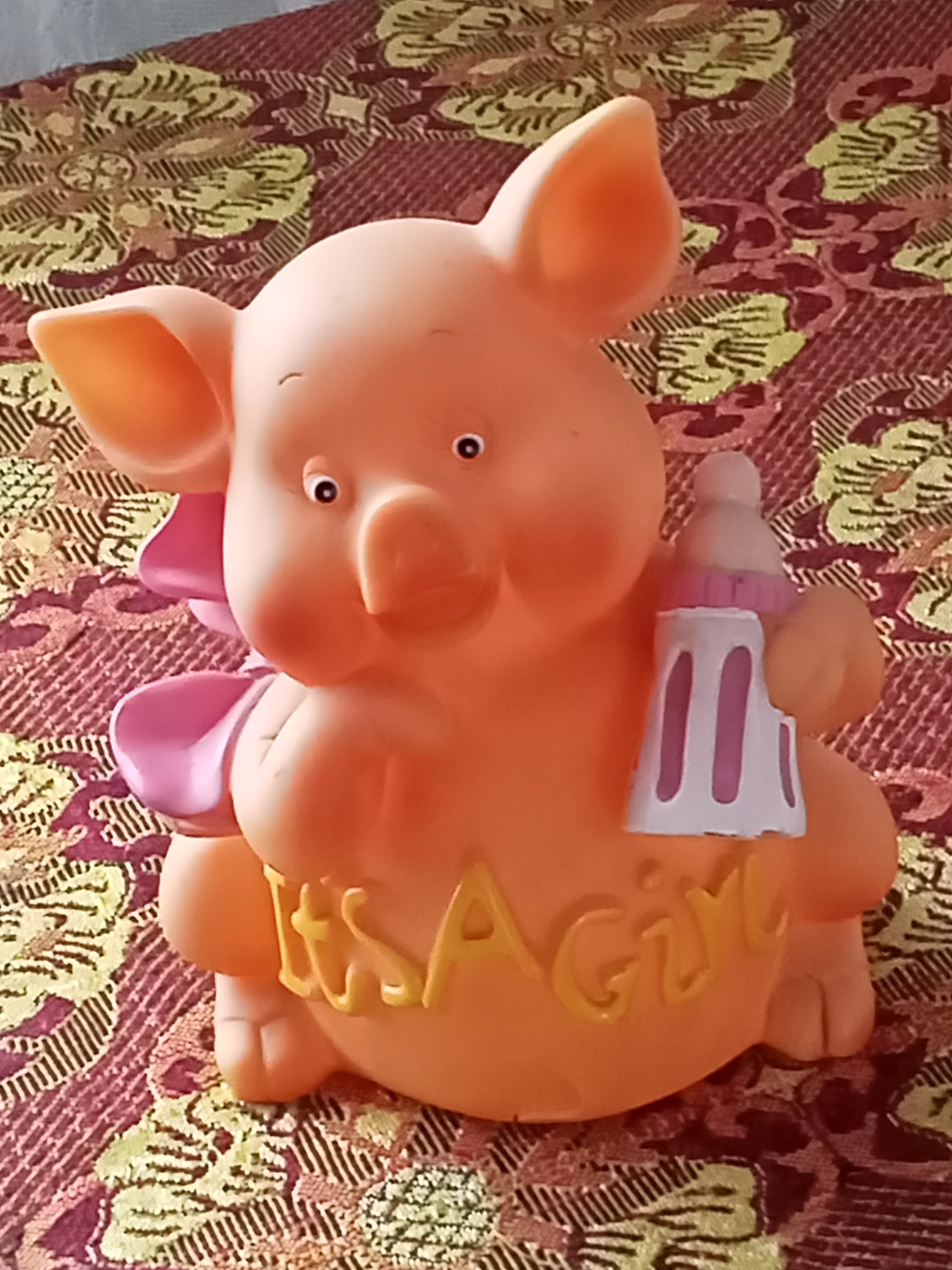 The image shows a close-up, eye-level view of an orange pig-shaped piggy bank sitting on a patterned fabric surface. The piggy bank is stylized with large ears, a snout, and painted eyes. It is holding a baby bottle in its right hand and has pink bows on its chest. The words "IT'S A GIRL" are written in yellow on the front of the piggy bank. The fabric surface has a repeating pattern of green and dark red floral or paisley designs. The lighting is moderate, and the image is in focus on the piggy bank. There is no information in the image that can be used to determine its location in Sokuluk, Kyrgyzstan.