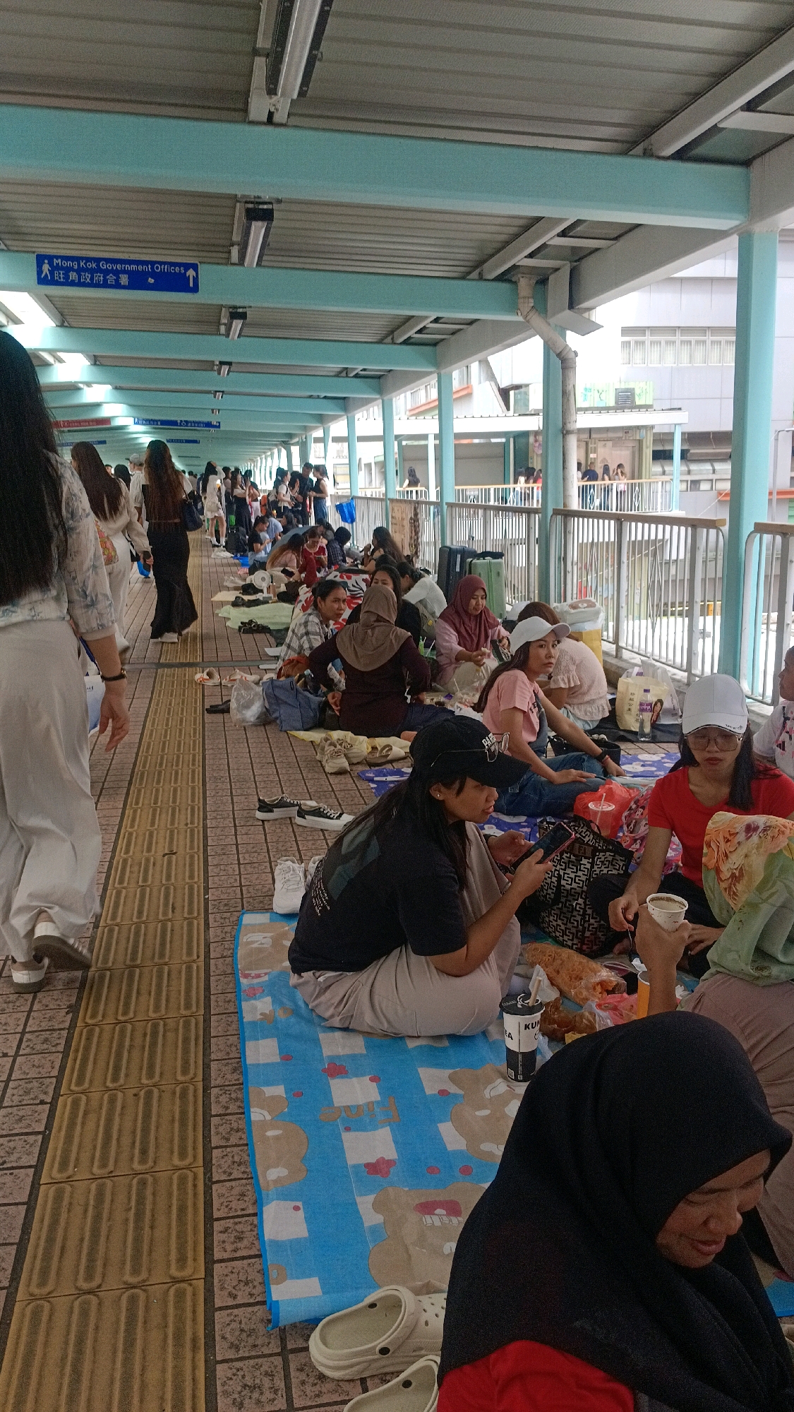In Hong Kong, China, a group of people are sitting on the ground in a covered walkway. The walkway has a light blue ceiling supported by white columns and a metal railing on the right side. On the left, there is a tactile paving walkway. Many individuals are seated on mats, with luggage and food items visible. Some are looking at their phones, while others are interacting with each other. The atmosphere appears relaxed, with people waiting or resting. A sign in English and Chinese indicates "Mong Kok Government Offices." The overall scene suggests a waiting area or a temporary gathering space.