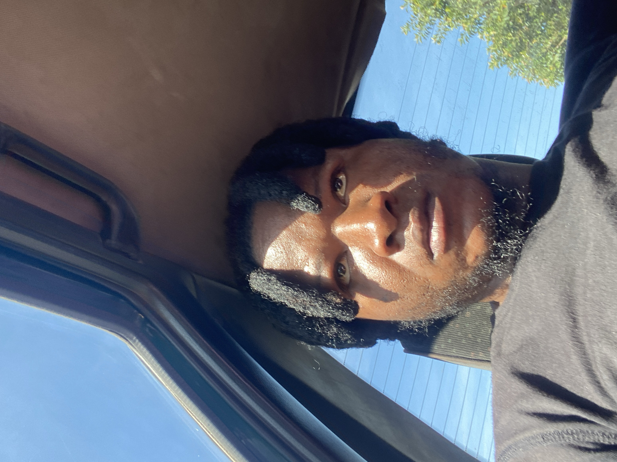The image is a close-up, selfie-style photograph of a Black man from the chest up, taken from inside a vehicle. The man has dark skin, black dreadlocks, and a short beard. He is wearing a black t-shirt. The interior of the car, possibly a sunroof or window frame, is visible at the top of the image. Sunlight illuminates the man's face from the side, creating strong shadows. Through the car window, a clear blue sky and green foliage are visible, suggesting an outdoor setting. The image is taken from a low angle, looking up at the man. The scene is likely set in Jos, Nigeria, given the context provided.