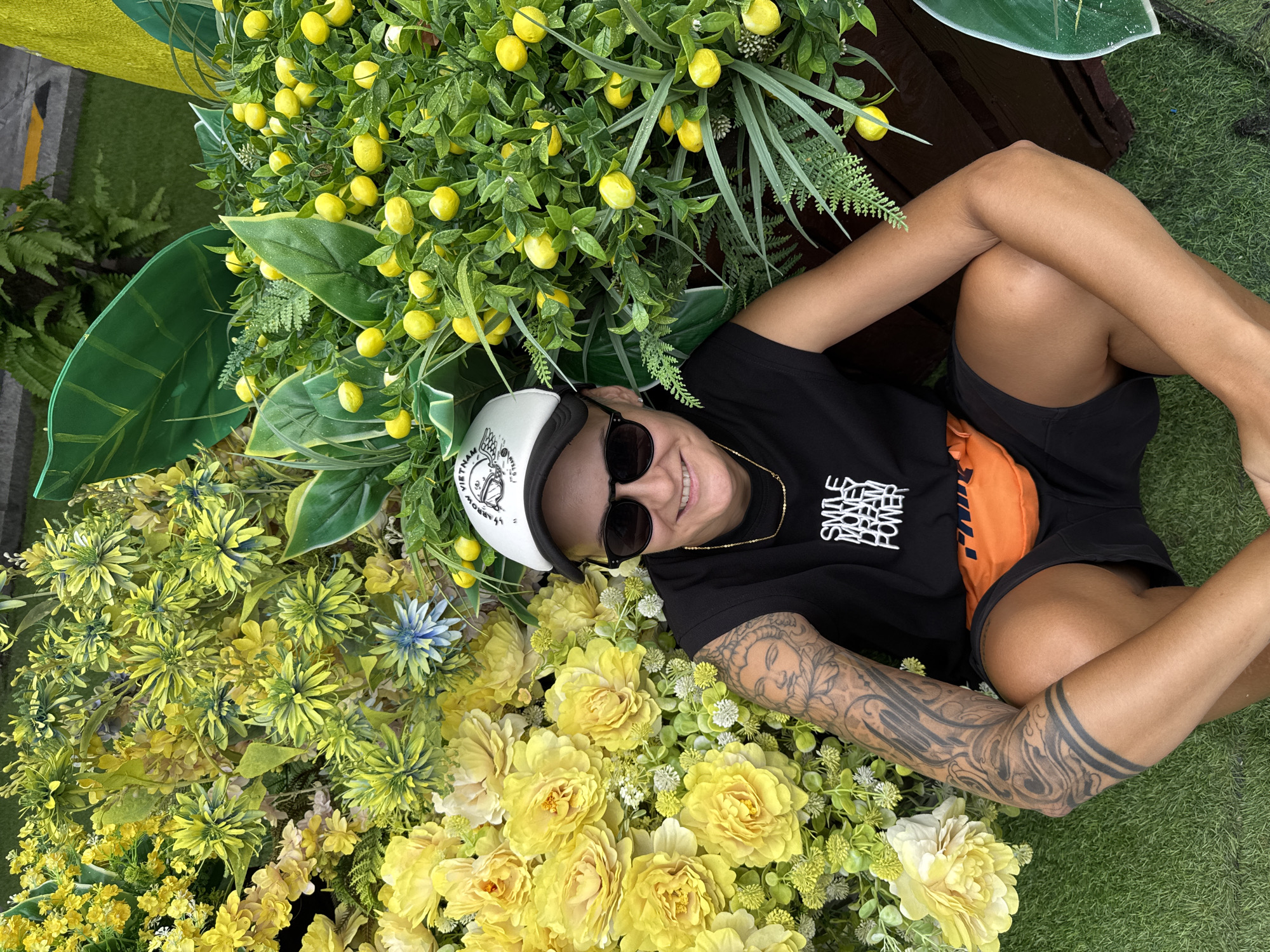A person is sitting on artificial grass in front of a floral and fruit-bearing display. The person is wearing a black t-shirt with white text that says "SMILE MONEY DREAM POWER," black shorts, an orange fanny pack, sunglasses, and a white and black trucker hat with an embroidered design. They have tattoos on their left arm and are smiling at the camera. The background features numerous artificial yellow flowers and artificial lemons on green foliage, with large green artificial leaves visible. The display is likely part of a temporary installation, possibly for an event or decoration in Nha Trang, Vietnam.