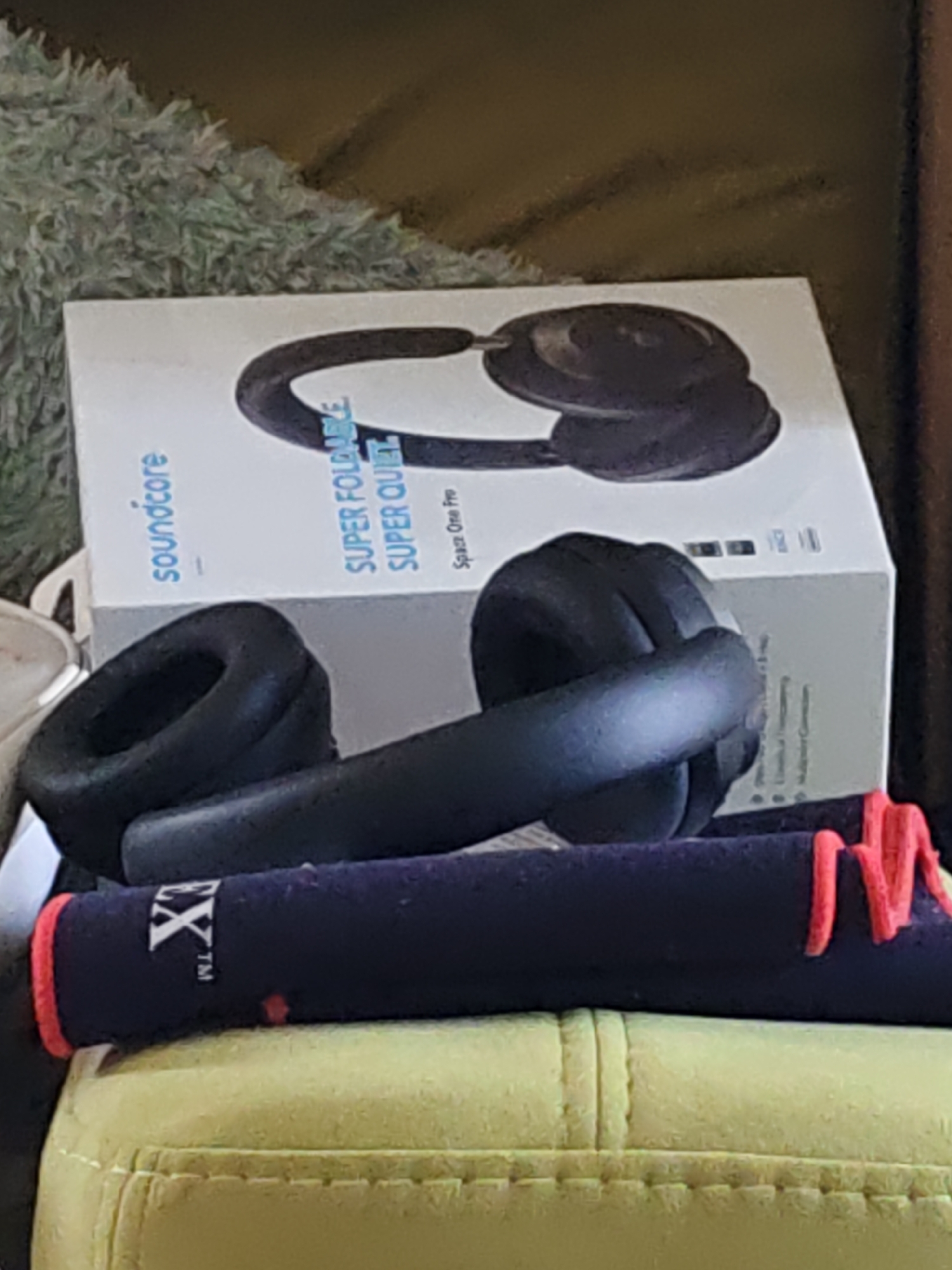 The image shows a pair of black over-ear headphones placed on top of a dark blue cylindrical object with red trim and white lettering that reads "EX". The headphones are in front of a white box with "soundcore" printed in blue on the left side and "SUPER FOLDABLE SUPER QUIET" in blue and black text on the right. The box also features an image of headphones. In the background, there is a textured green surface. The scene appears to be indoors, on a surface that looks like a couch or upholstered furniture. The location is not specified within the image, and therefore the city of Rostov-on-Don, Russia, cannot be inferred from the visual information provided.