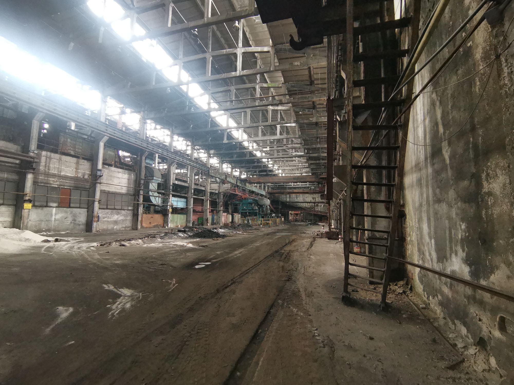 The image depicts the interior of a large, abandoned industrial building in Zhivkovo, Bulgaria. The space is characterized by high ceilings with exposed metal beams and skylights allowing daylight to enter. The floor is dirt and debris-covered, with visible tracks suggesting the movement of heavy machinery. In the foreground, a rusty metal ladder is attached to a heavily weathered wall. Further into the space, industrial equipment and machinery can be seen. The overall environment suggests decay and disuse. There are no people visible in the image.