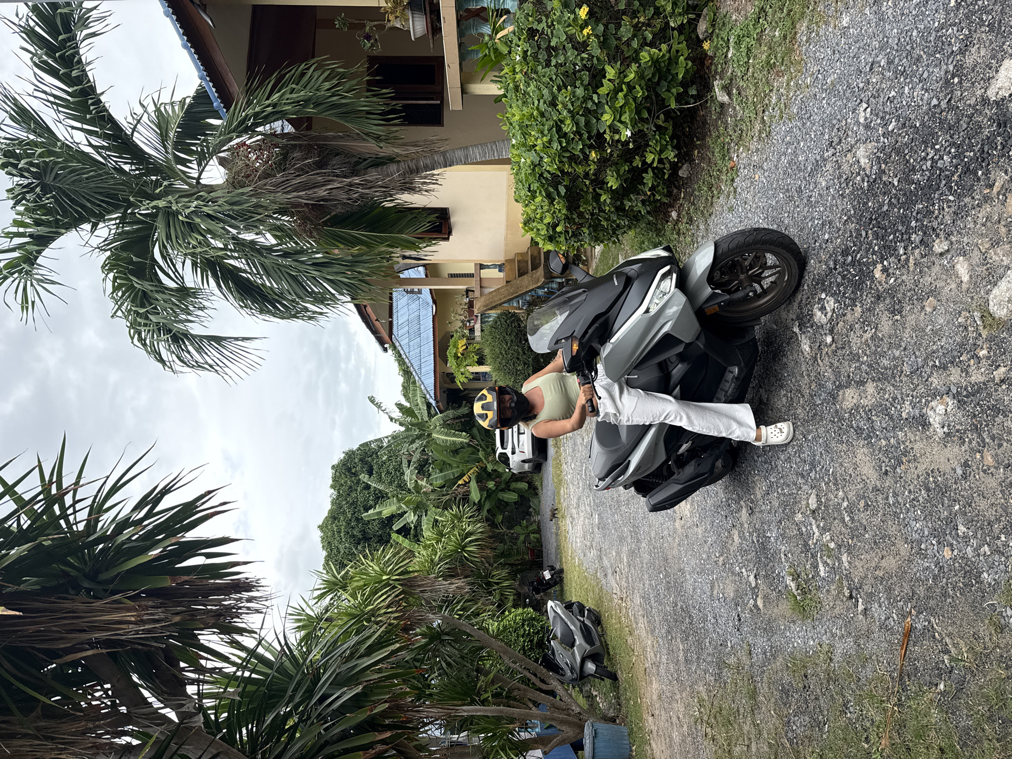 A woman wearing a green tank top, white pants, and a yellow and black helmet is sitting on a silver and black scooter in a gravel driveway. She is wearing white clogs with an animal design. The scooter is facing forward, with its headlights on. Behind her, there is a white SUV parked, a black scooter, a blue trash can, and a cat walking in the driveway. The environment is tropical, with palm trees and lush green bushes. Two houses with blue and brown roofs are visible in the background. The sky is overcast. The image appears to be taken in the Mae Nam area of Ko Samui District, Thailand, known for its residential properties and local transport.