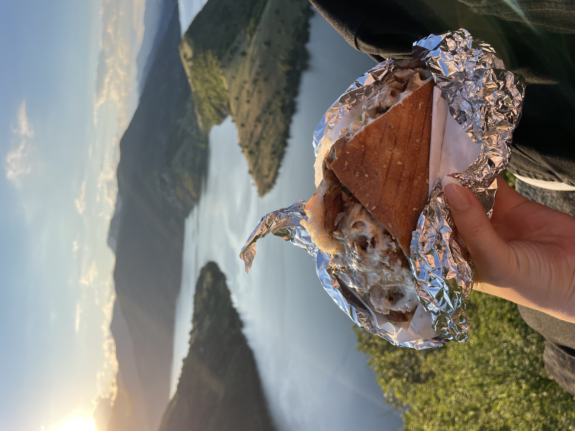 In the foreground of the image, a hand holds a sandwich wrapped in aluminum foil. The sandwich appears to be cut in half, revealing layers of filling and sauce. The background depicts a picturesque landscape with a large body of water winding through hills and mountains. The sun is setting, casting a warm glow over the scene. The location is identified as Borovitsa, Bulgaria, suggesting a scenic overlook or a viewpoint near the Arda River. The overall impression is one of enjoying a meal amidst a beautiful natural setting.