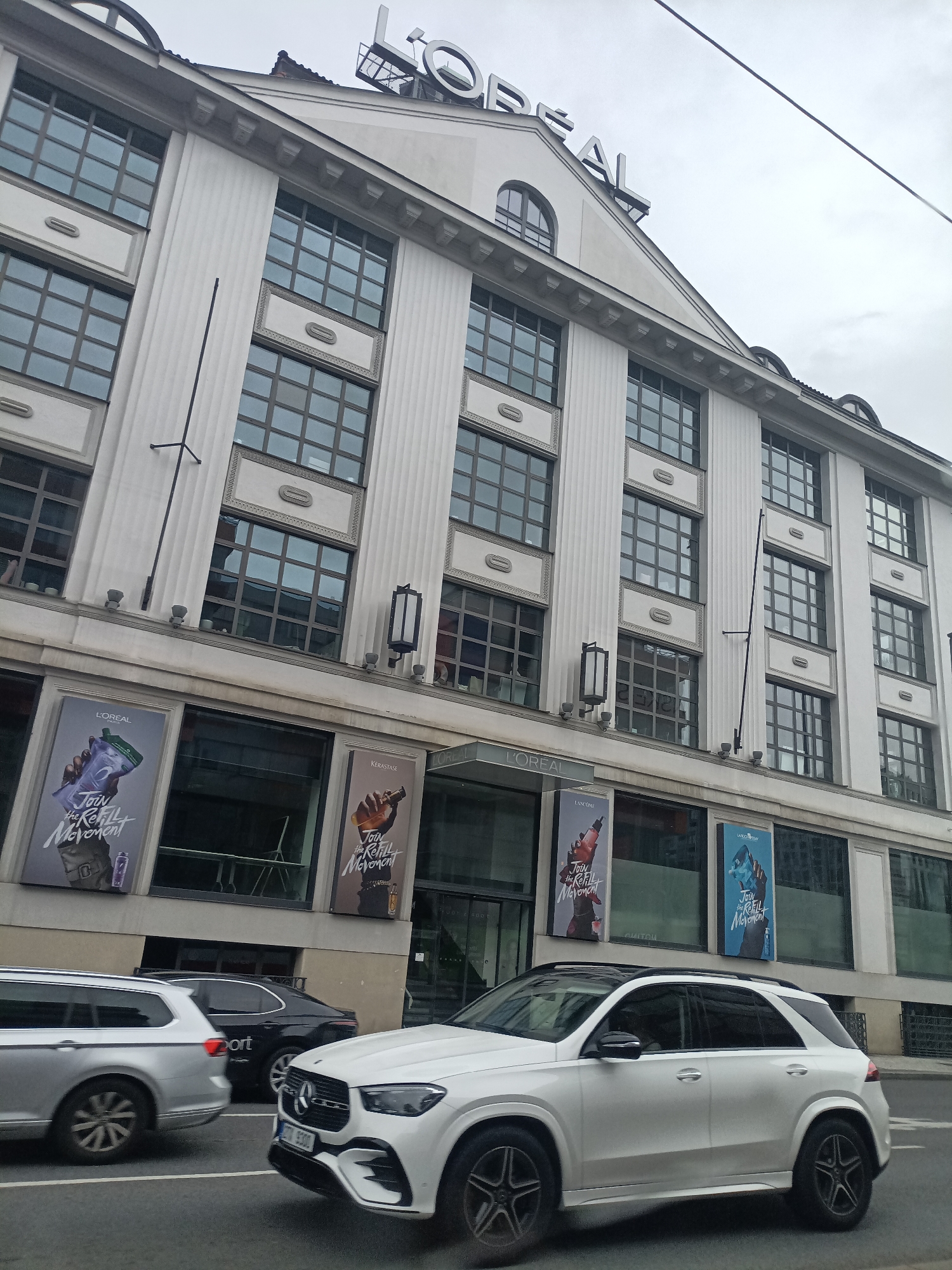 The image shows the L'Oréal building, located at Pařížská 18, Prague, Czechia. The building is a multi-story white structure with many windows. Large L'Oréal advertisements are displayed on the lower level. In the foreground, a white Mercedes-Benz SUV is driving on the street, and to its left, a silver station wagon is visible. The sky is overcast.