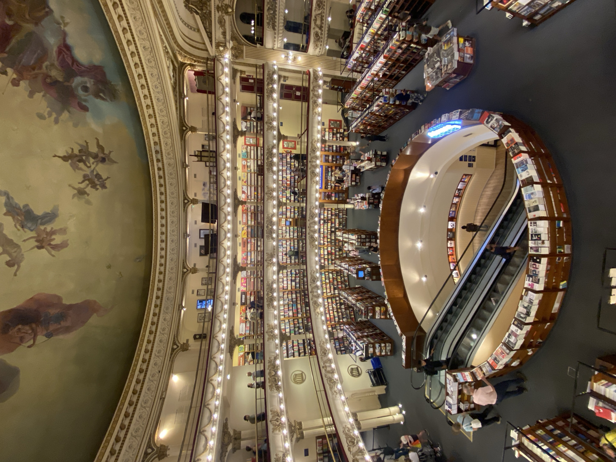 The image depicts an expansive interior view of the El Ateneo Grand Splendid bookstore, located in Buenos Aires, Argentina. The scene is a multi-level circular structure, a converted theater, observed from an elevated perspective. A large, ornate domed ceiling dominates the upper frame, featuring classical frescoes with numerous figures, including cherubs and allegorical subjects, painted in muted reds, blues, and browns against a light greenish-gold background with cloud formations.
Below the ceiling, a series of at least three decorative balcony levels with gilded railings and integrated lighting encircle the central space. Each balcony is lined with floor-to-ceiling wooden bookshelves densely packed with books. The main ground floor is dark-colored, likely carpeted, and features additional freestanding wooden bookshelves and display tables arranged throughout.
In the mid-ground, a prominent set of parallel escalators descends into a lower, unseen level. These escalators are partially encircled by a curvilinear wooden display counter exhibiting numerous books. Scattered throughout all visible levels are approximately 30-40 casually dressed individuals of varying ages. People are observed browsing bookshelves, walking along aisles, and riding the escalators. Specific signage, including "MANGA," is visible on an upper-level shelf. The entire space is illuminated by warm artificial lighting emanating from fixtures along the railings and recessed in the ceilings.