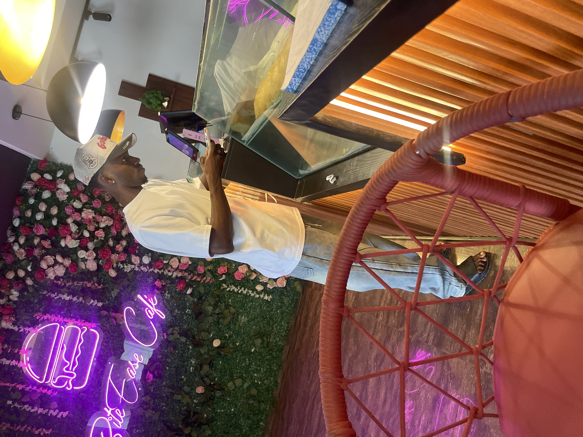 A man stands in what appears to be a cafe in Adankolo, Nigeria. He is light-skinned, wearing a white t-shirt, jeans, and a baseball cap. He is holding a device, possibly a tablet or a phone, and looking at it. Behind him is a wall decorated with flowers and a neon sign reading "Bite Tease Cafe" with a burger icon. To his right is a partially visible red chair and a structure that looks like a display case. The cafe has a wooden ceiling. The lighting suggests it is either evening or the interior is dimly lit.