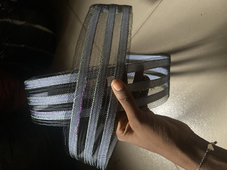 A hand with dark skin holds a crafted object of stacked, striped mesh. The mesh is black with metallic silver and gold lines. The object's shape suggests a container or headpiece, constructed from layered loops of the mesh. The hand has a simple silver bracelet with small heart charms. The background shows a shiny black surface and a lighter, tile-like surface.