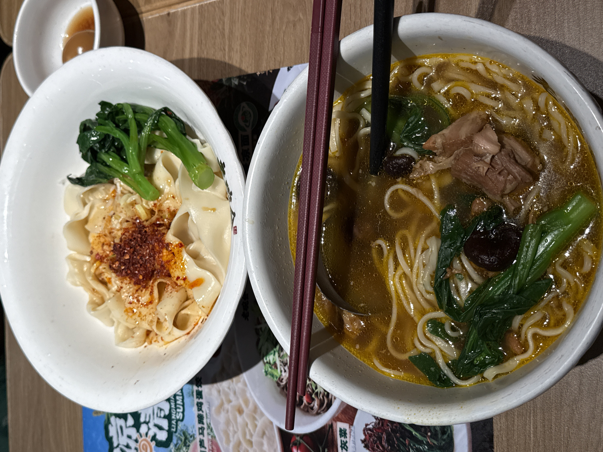 In the Futian District, China, two bowls of noodles are presented on a wooden table. The bowl in the foreground contains a rich, golden broth filled with white noodles, green leafy vegetables, chunks of meat, and a dark mushroom. A ladle rests in the broth, and a pair of black chopsticks is positioned across the bowl. The bowl in the background holds a portion of noodles with chili flakes and blanched green vegetables, accompanied by a small dish of dipping sauce to the right. The scene is likely a restaurant setting.