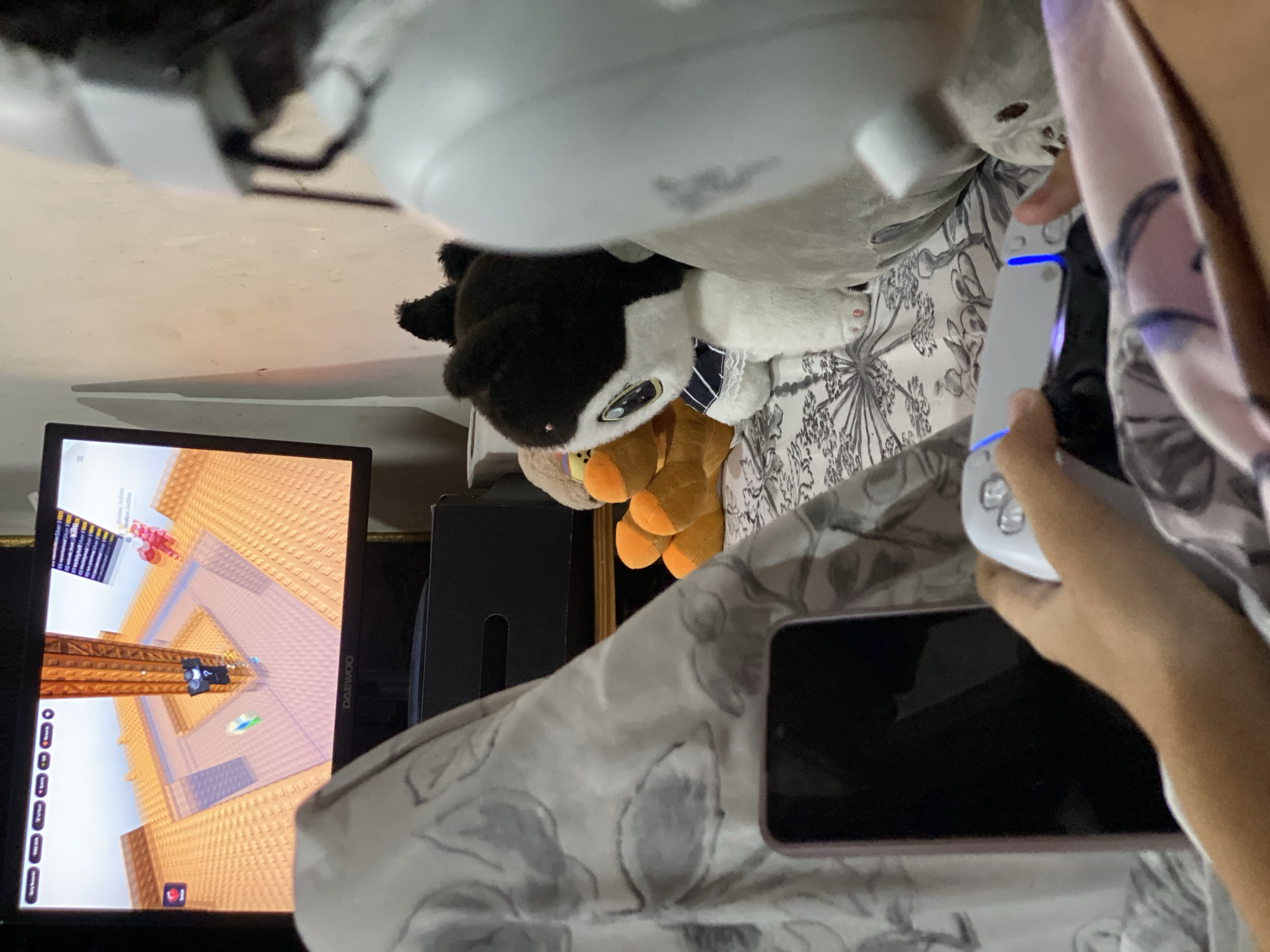 The image shows a person holding a white PlayStation 5 controller, seemingly playing a game on a monitor displaying a Roblox game with an obstacle course. The monitor is a black, modern flat screen. Next to the monitor, there appears to be a black electronic device, possibly a PlayStation 5 console, although its shape is partially obscured. A black and white plush toy lies on a patterned bedding, and an out-of-focus phone is partially visible in the foreground. There is a large white object hovering over the monitor that appears to be an upside-down ceiling fan, obscuring the upper view of the image. The location is indeterminable based solely on the visible objects and surroundings; the image contains no information about Buenos Aires, Argentina.