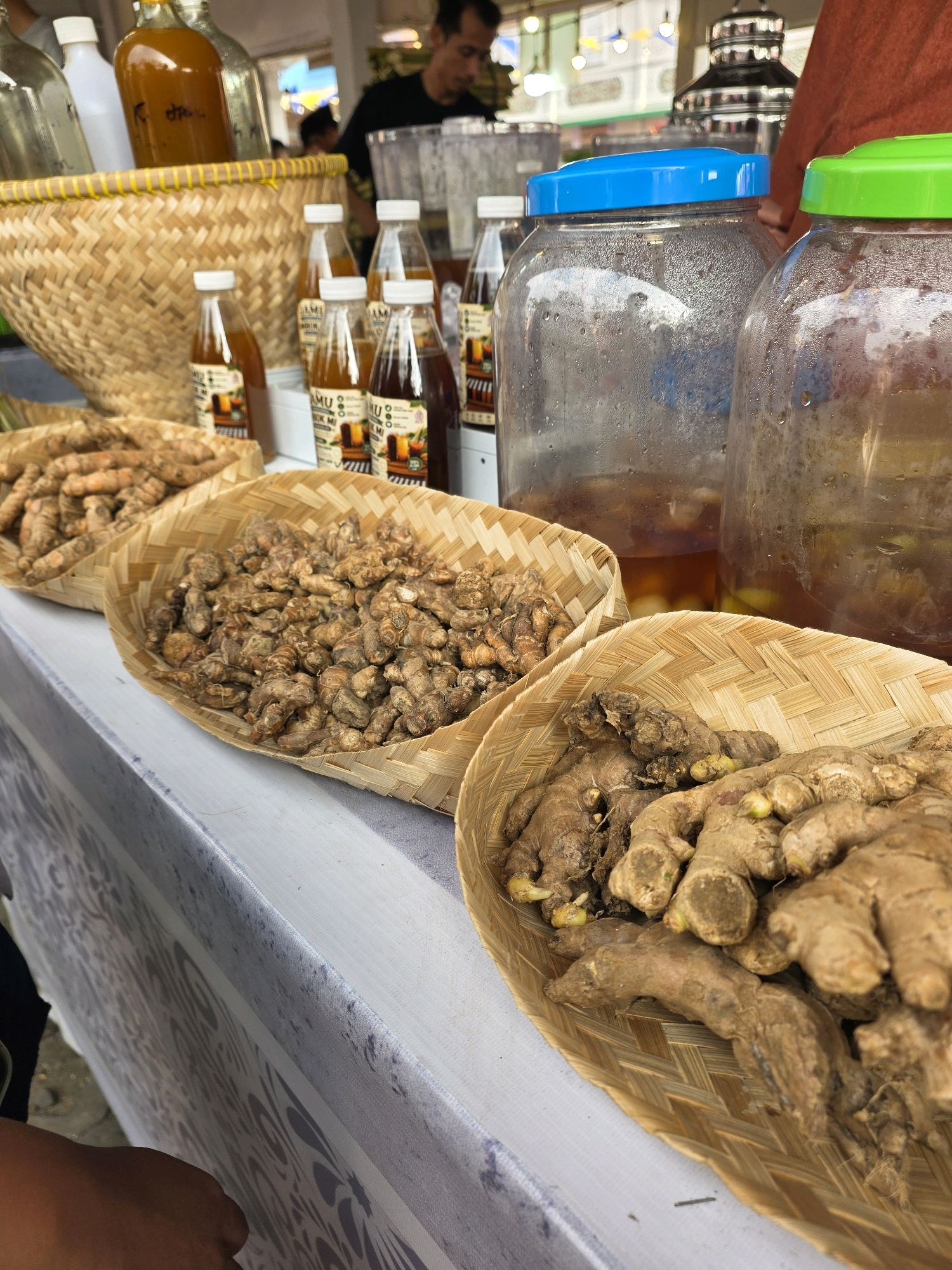 This image depicts a market stall, likely in Bandung City, Indonesia, showcasing a variety of traditional Indonesian herbal drinks, known as "jamu," and fresh ginger and turmeric roots. In the foreground, three woven bamboo baskets are filled with these roots. The basket on the right contains larger, fresh ginger pieces, while the middle basket holds smaller, dried turmeric or ginger pieces. The basket on the left is partially visible and also contains dried roots. Behind these baskets are several small glass bottles with white caps, filled with amber-colored liquids and labeled "JAMU." To the right of the bottles are two large glass jars with blue and green lids, respectively, containing a dark amber liquid, suggesting they are for storing or serving jamu. In the background, a man in a black t-shirt is visible, appearing to be tending to the stall. The stall is set up on a table covered with a white cloth featuring a grey patterned design. The overall setting suggests an outdoor market environment, with hints of a bustling urban atmosphere in the blurred background.