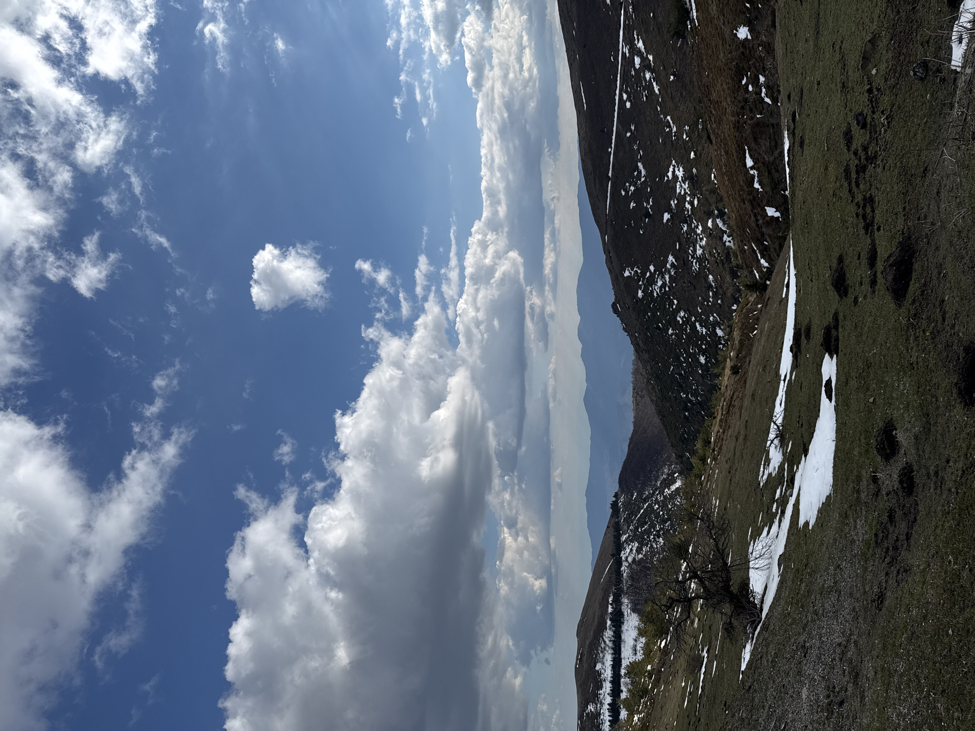 The image shows a mountainous landscape in Beli Osam, Bulgaria. The sky is mostly blue with scattered white clouds. A mountain slope covers the right side of the frame, displaying green grass mixed with patches of melting snow. Higher up the slope, the vegetation is sparser and darker. In the distance, layers of mountains are visible, fading into a hazy blue. A bare tree sits on the slope in the foreground.