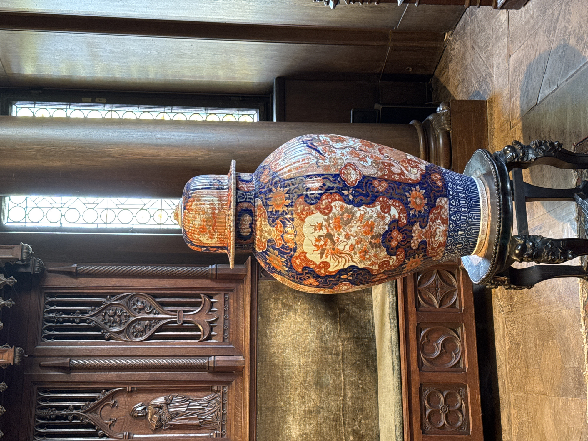 A large, ornate vase sits atop a dark wood stand within a room featuring detailed wood paneling and a window with a grid pattern. The vase has a globular body with an extended neck, and is decorated with intricate orange, white, and blue designs. The wood paneling behind the vase is dark and features carved elements. The wood of the floor and support pillar, visible on the right, suggests a historical location, likely the Cochem Castle in Cochem, Germany.