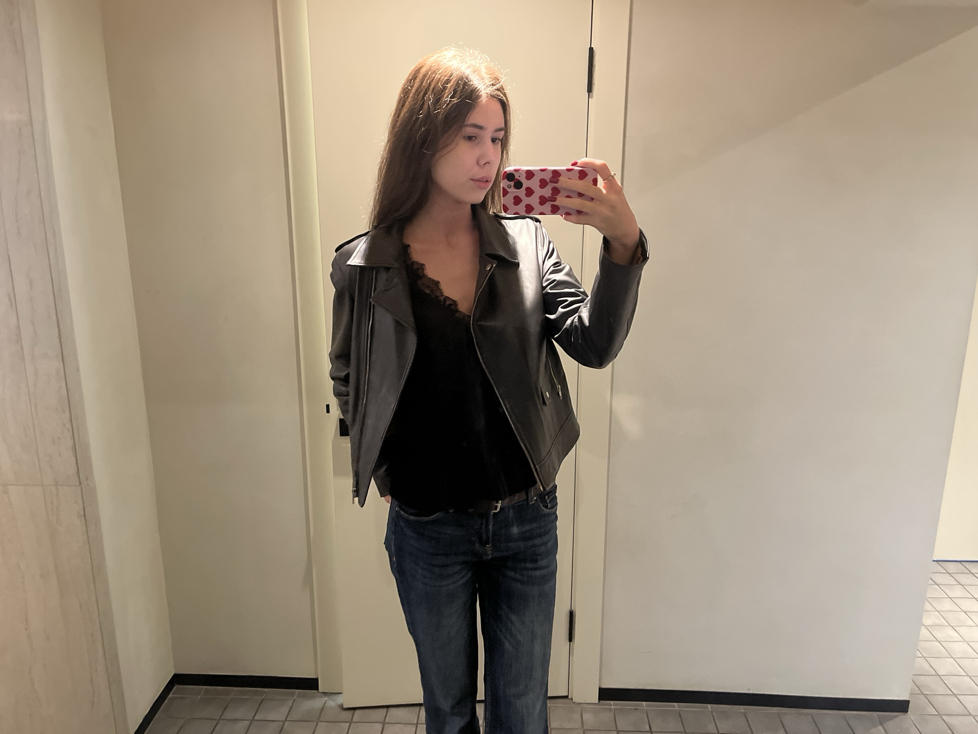 A person stands in a room and looks at the camera, taking a picture of themselves. The person has long brown hair and wears a dark-colored top with a black leather jacket and dark-wash jeans. They hold a cell phone in their right hand with a pink case decorated with red hearts. The environment appears to be a dressing room or bathroom, suggested by the neutral-colored walls and a tiled floor visible at the bottom right of the frame. The lighting in the room seems artificial, and the details in the background are somewhat blurred.