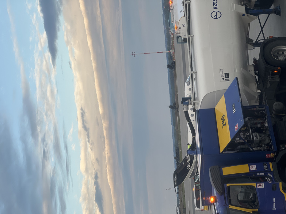 The scene takes place at Rzeszow-Jasionka Airport in Jasionka, Poland (latitude: 50.114, longitude: 22.026). A fuel truck is parked in front of a private jet, marked with the registration "OK-ESC." The truck has a blue and yellow paint scheme with the number "290" displayed on its side. The truck's fuel tank is white and bears the logo of "Rzeszow Airport" and a smaller logo of "LOT," a Polish airline. The truck's fuel tank is partially open, revealing a mechanical apparatus and a red warning light. The jet is partially visible behind the fuel truck. The background is dominated by a clear sky with white clouds. A red and white airport light tower is visible in the distance. The airport tarmac is paved and there are green trees and grass visible in the distance.