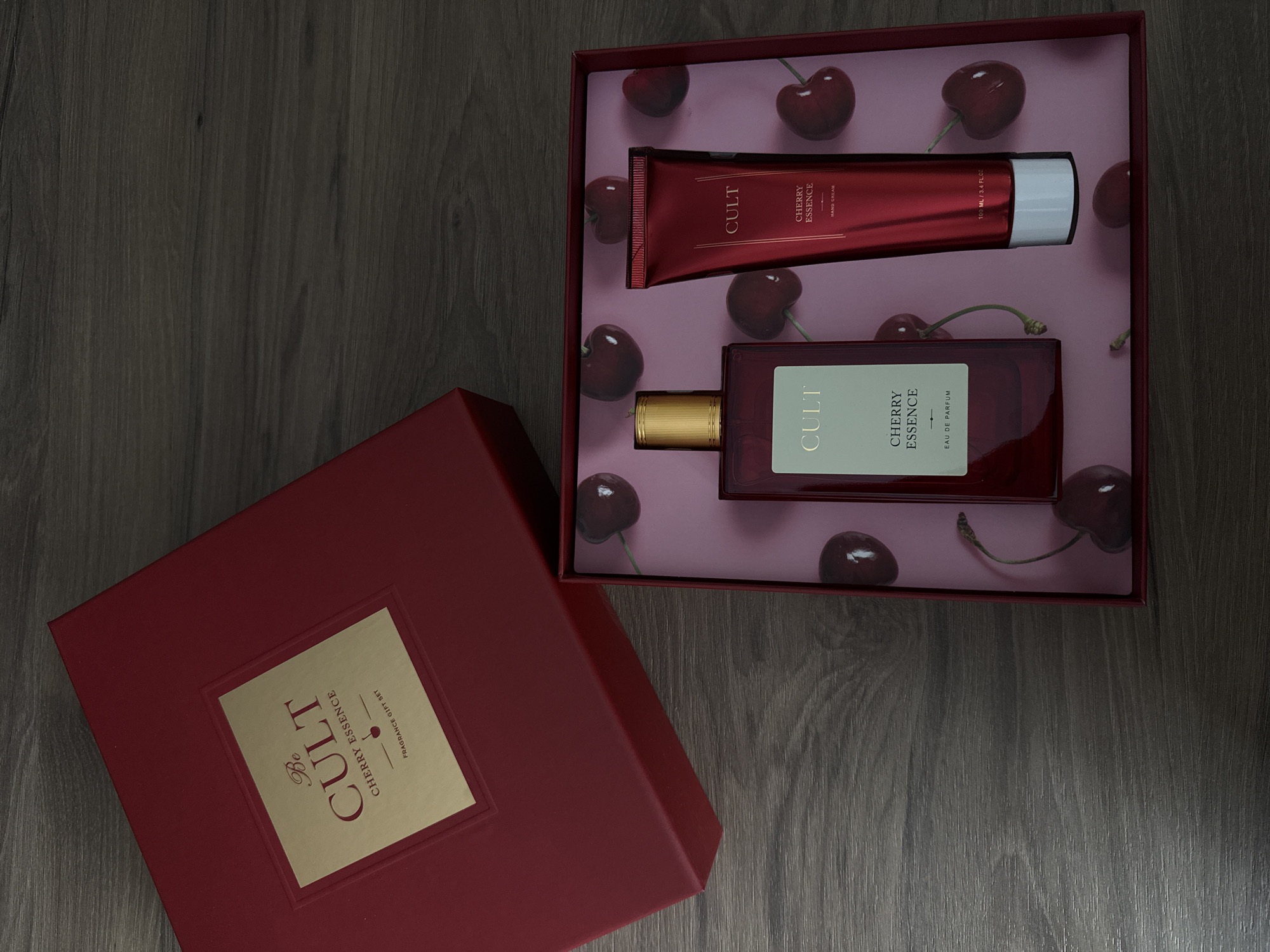 A red gift box for a fragrance set is open, revealing a perfume bottle and a tube of hand cream. The interior of the box is decorated with a pink background featuring a pattern of cherries. The perfume bottle is dark red glass with a gold cap and a label that reads "CULT CHERRY ESSENCE EAU DE PARFUM". The hand cream tube is also red and has a label that reads "CULT CHERRY ESSENCE HAND CREAM". The gift box itself is dark red and has a gold label on the lid with the text "Be CULT CHERRY ESSENCE FRAGRANCE GIFT SET". The items are arranged on a dark wooden surface. There are no people or animals present in the image. The location in the city of Murom, Russia is not discernible from the image.