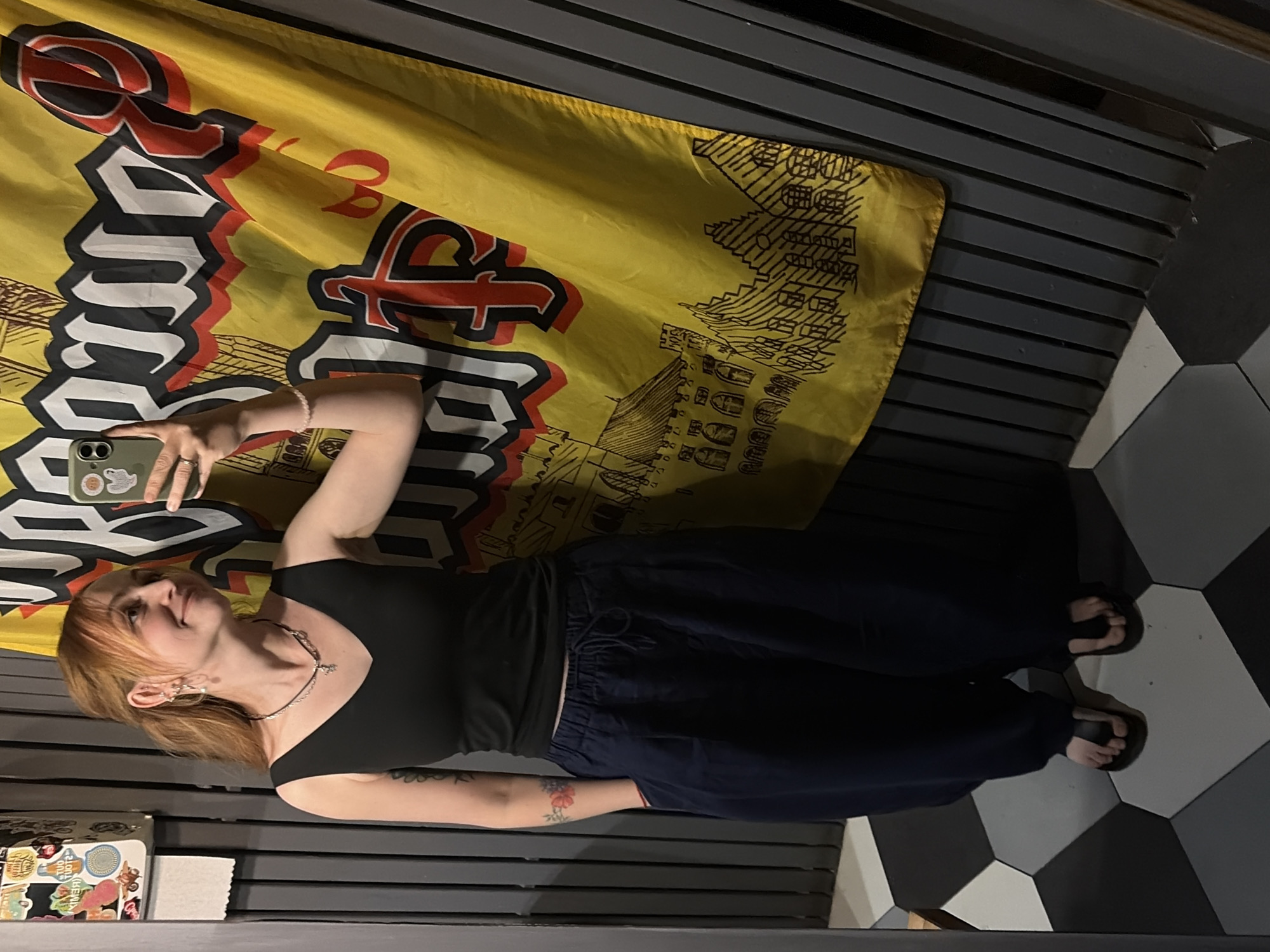 A woman stands in an interior space, taking a selfie with a smartphone. She is wearing a black top and dark, loose-fitting pants. A large, yellow flag with bold, stylized writing and graphics hangs behind her, partially covering the wall. The background includes a gray, slatted wall and a black-and-white hexagonal tiled floor. A small table or shelf with stickers and decorative items is visible in the lower left corner of the image. While the image does not give specific details about the city of Chaikovsky, Russia, or its precise location, the presence of the flag suggests a location within a specific establishment or venue that could be tied to that city, but without further information, the conclusion is limited.