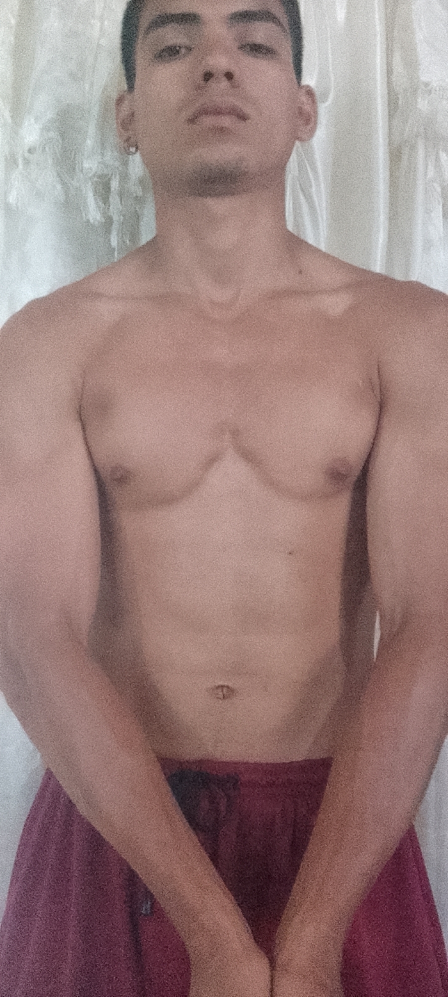A single male individual is depicted in a front-facing, waist-up portrait, located indoors in the city of Juan Griego, Venezuela. The subject is shirtless, displaying the chest and abdomen, and wears maroon-colored shorts. The individual's arms are positioned in front of the lower abdomen, with forearms angled downwards and hands clasped. The subject has short dark hair and minor facial stubble. A silver-colored hoop earring is present in the left earlobe. The background is a plain, light-colored surface with subtle vertical textures, resembling a wall or curtain. The image displays a grainy texture.