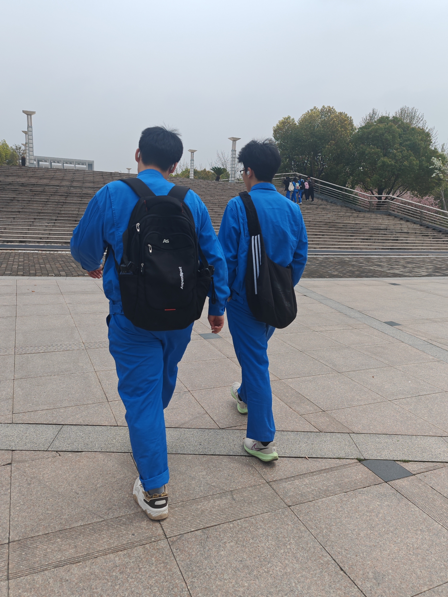 Two people wearing matching blue jumpsuits walk away from the viewer on a paved surface in Jiangning District, China. The person on the left wears a black backpack with the word "AspenSport" written on it, and white sneakers. The person on the right carries a black Adidas side bag and wears light green sneakers. Behind them are concrete stairs leading up to a plaza with decorative light posts, trees, and other people. The sky is overcast.