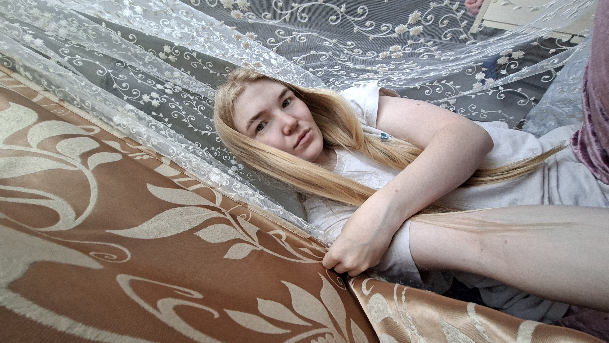 A fair-skinned female with long blonde hair is reclining on what appears to be a bed, partially obscured by patterned fabrics. She is wearing a light-colored t-shirt. The fabrics include a semi-transparent white floral patterned fabric and a brown fabric with a large leaf pattern. The environment suggests an interior setting, possibly a domestic space. There are no indications of the city of Большой Букор, Russia.