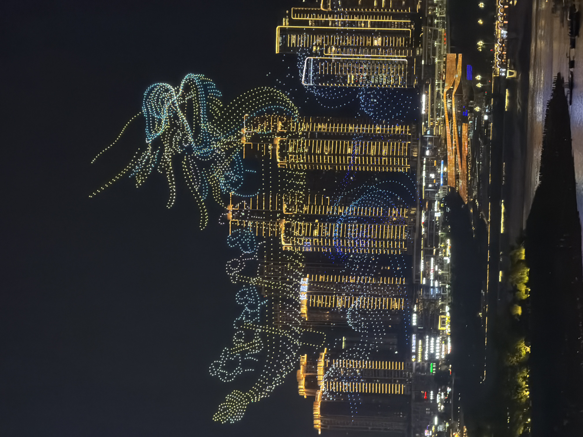 This media file captures a nighttime drone show in Yuzhong District, China. The primary subjects are a formation of numerous drones in the sky, creating luminous patterns against the dark backdrop. These drones are illuminated with yellow and blue lights, forming intricate shapes that appear to depict a dragon or similar mythological creature with flowing tendrils.
The scene also includes a cityscape with tall buildings illuminated by countless yellow and white lights, suggesting a densely populated urban environment. These buildings are arranged in rows and exhibit a grid-like structure. A body of water, likely a river, is visible to the right of the cityscape, reflecting the lights from the buildings. Trees and possibly a waterfront promenade can also be discerned along the water's edge, also illuminated with yellow lights.
The event taking place is the drone show, a spectacular display of aerial light art. The time of day is clearly night, indicated by the dark sky and the prominence of artificial lighting. The weather appears clear, allowing for the drone performance. The location cues of the cityscape, the river, and the specific district point to an urban setting within China. There is no visible text in the image. The overall impression is one of a grand, celebratory, or artistic event, likely intended to impress and entertain.
