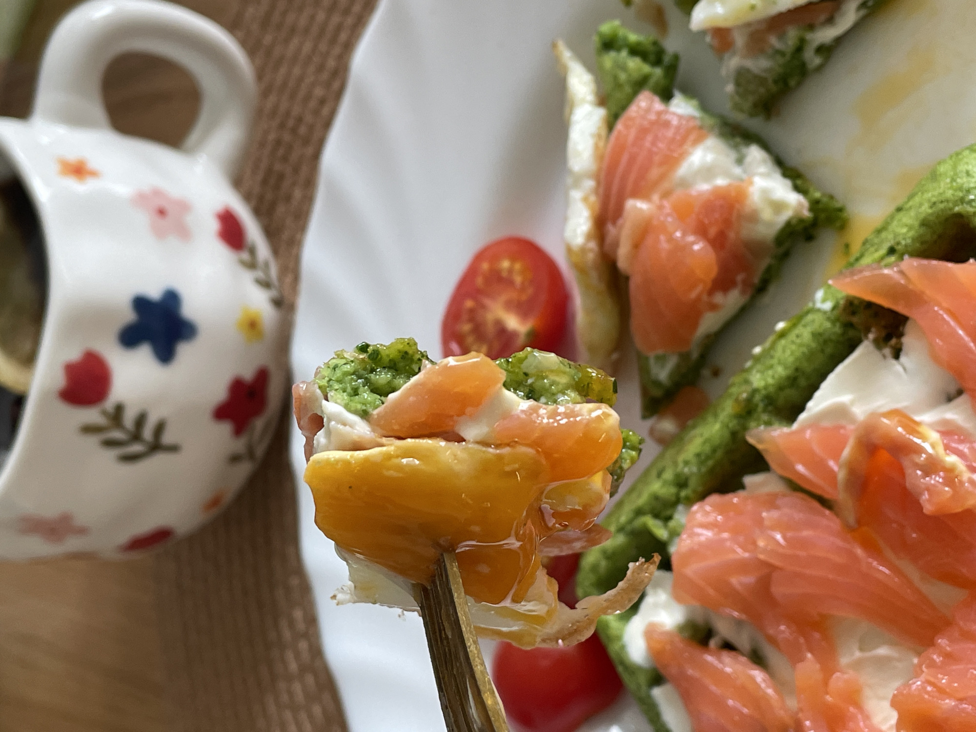 The image shows a plate of green pancakes topped with cream cheese and smoked salmon, along with cherry tomatoes. A white mug with a floral pattern is visible in the background, sitting on a brown surface. One of the pancakes is held up with a fork, revealing a layer of fried egg beneath the salmon. The city of Chaikovsky, Russia, is not discernible from this image.