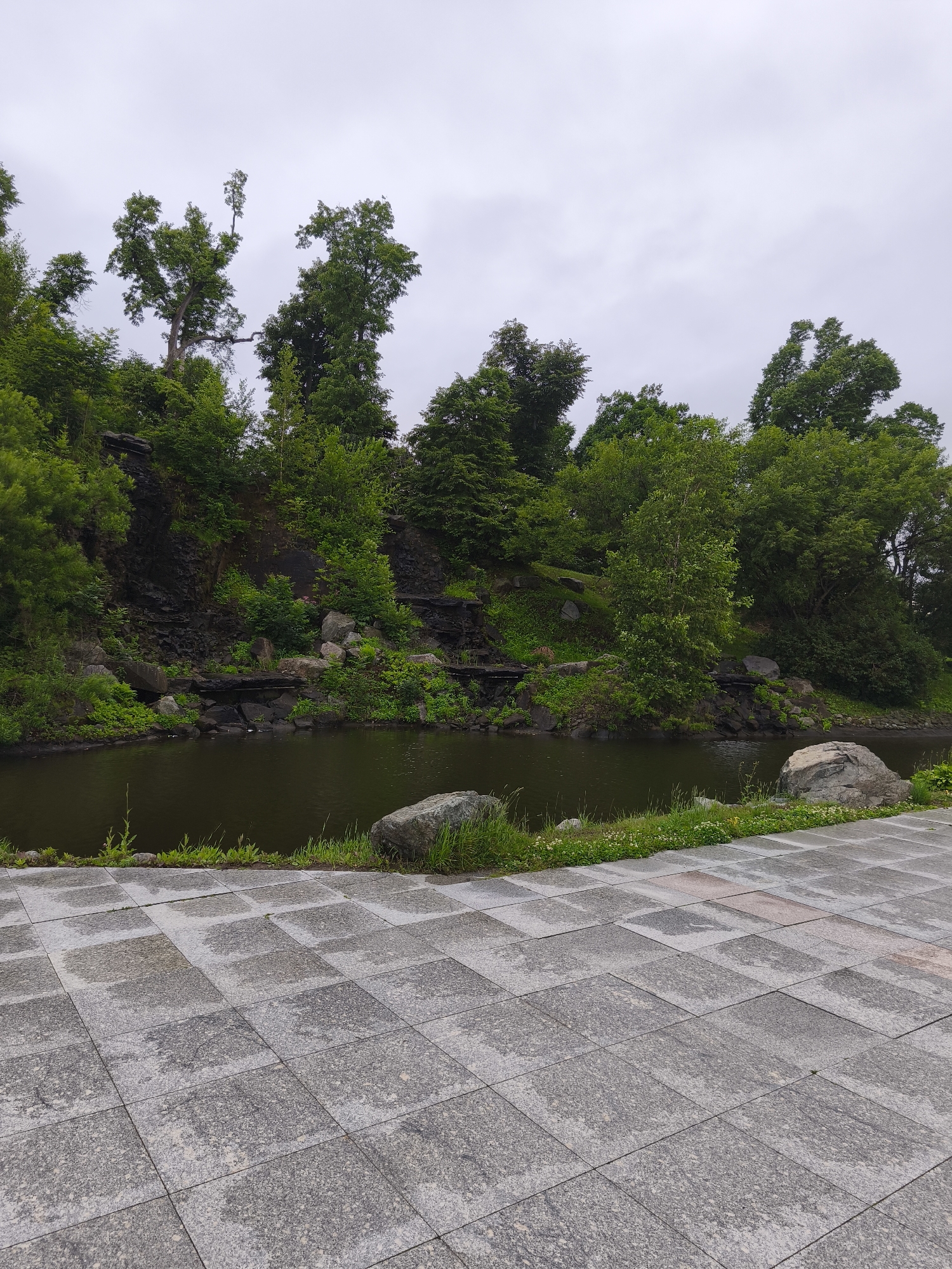 The image shows a park-like setting with a body of dark water on the left, surrounded by lush green trees and rocky terrain. The foreground is a paved area with large, square gray tiles. The sky is overcast and gray. In the middle ground, the water reflects the dense foliage of trees and bushes growing on a rocky incline. Some rocks are visible near the water's edge. The background consists of more trees and a hill. The overall impression is of a natural landscape within a developed area, possibly a park or botanical garden. There are no people or animals visible in the image.