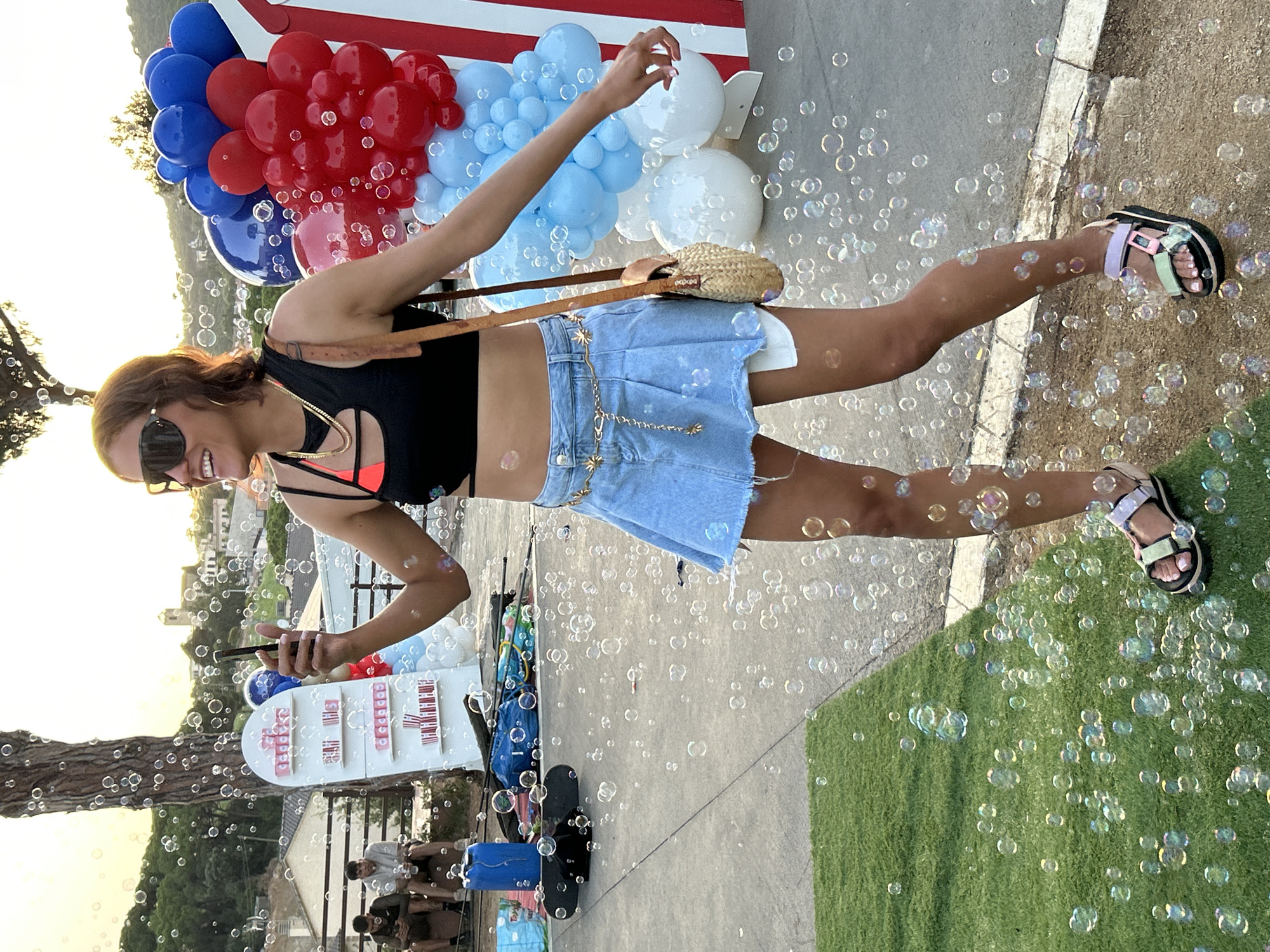In Sant Vicenç De Montalt, Spain, a woman with medium-length brown hair and sunglasses stands outdoors, holding a black smartphone in her right hand. She is wearing a black cutout sports bra with pink accents, a light-wash denim mini-skirt with a distressed hem, and a decorative gold chain belt with star charms. She has a small woven crossbody bag with a brown leather strap. Her left arm is extended slightly to her side, and she is wearing platform sandals with pastel-colored straps. Numerous bubbles float around her, obscuring parts of the background.
To her left, two men are seated on a bench. The man on the left wears a dark t-shirt and dark shorts, while the man on the right wears a grey t-shirt and khaki shorts. Both are looking at their phones. Behind them, a white shelf unit displays multiple rows of pink cups and red and white striped containers.
In the background, there is a tall tree on the left, and a cluster of red, blue, and white balloons to the right, with white and red striped fabric behind them. The setting appears to be a paved area next to a patch of artificial turf, with a slight incline leading to buildings and a hill in the distance, suggesting a town or village. The lighting indicates it is late afternoon or early evening.
