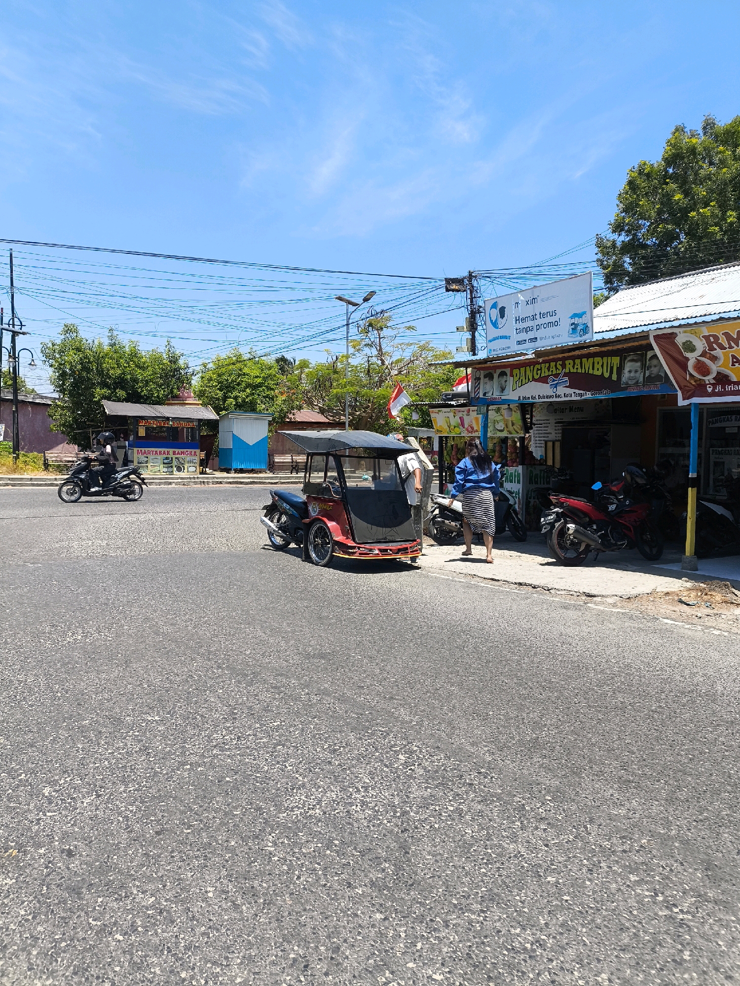 The image captures a street scene in Gorontalo, Indonesia, on a clear, sunny day. In the foreground, a paved road takes up a significant portion of the frame. A red and black three-wheeled motorcycle, commonly known as a becak motor, is parked on the right side of the road, partially obscuring a row of shops.
Several other motorcycles are also visible. One black motorcycle with a rider wearing a helmet is moving on the road to the left. Another red motorcycle is parked in front of a shop.
The shops themselves are colorful, with awnings and various signs. One prominent sign above a shop reads "PANGKAS RAMBUT," indicating a barbershop. Another sign, a large banner hanging from a pole, displays text in Indonesian and features an illustration of a blue vehicle and two figures wearing face masks, promoting a promotion related to "hemat terus tanpa promo!"
A woman in a blue shirt and a striped skirt is standing on the sidewalk near the shops, facing away from the camera. Another person, partially visible, stands near the becak motor. Behind the shops, trees and power lines are visible against a bright blue sky with sparse white clouds. The overall scene suggests a bustling urban environment with local businesses and transportation.