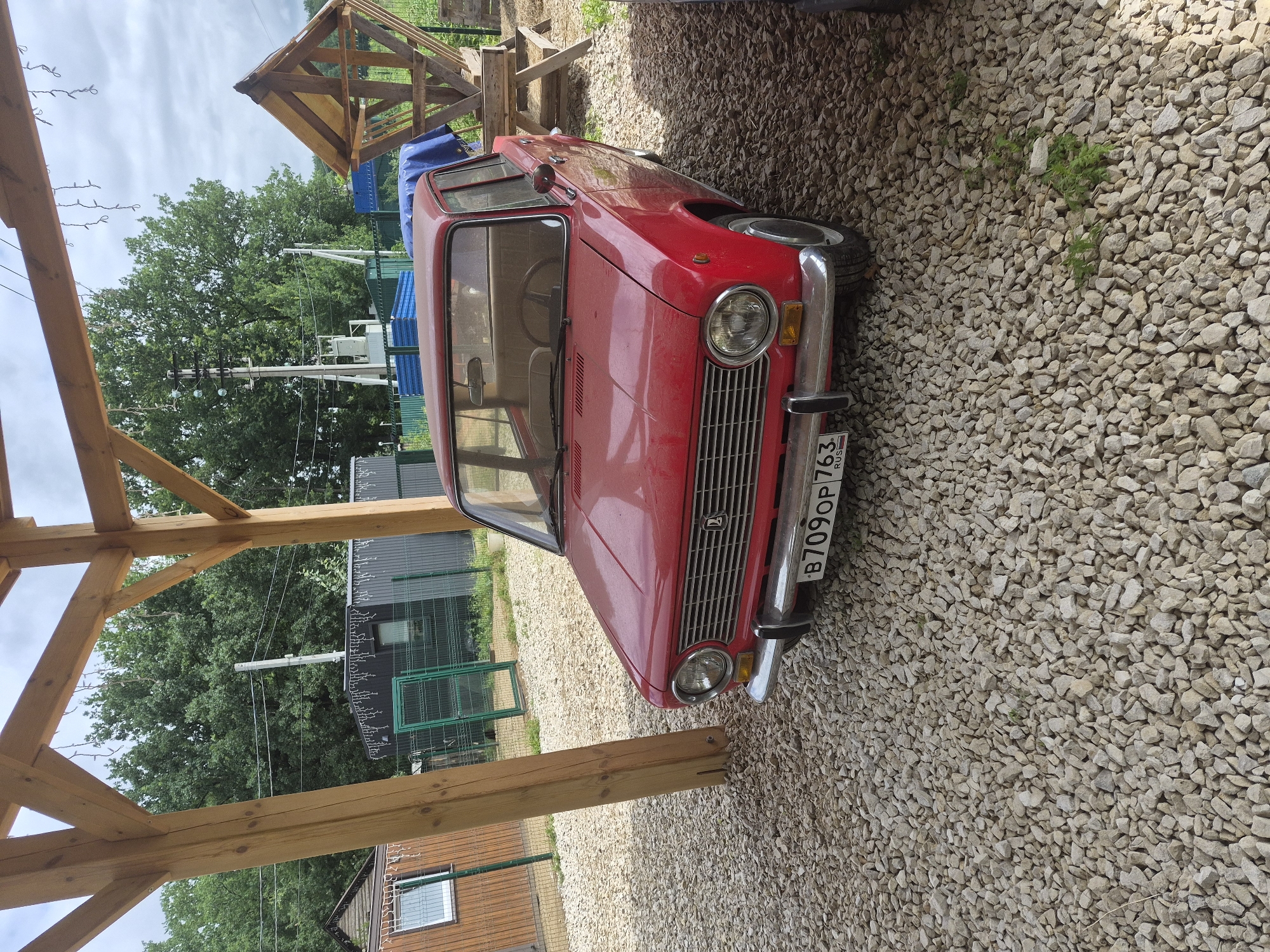A red Lada car is parked on a gravel lot under a wooden structure with a thatched roof. The car is facing forward and is slightly to the right of the center of the image. The car's license plate reads "B709OP 763 RUS". In the background, there are trees, a fence, and some buildings. The sky is overcast. The scene appears to be in the city of Volzhskii, Russia.