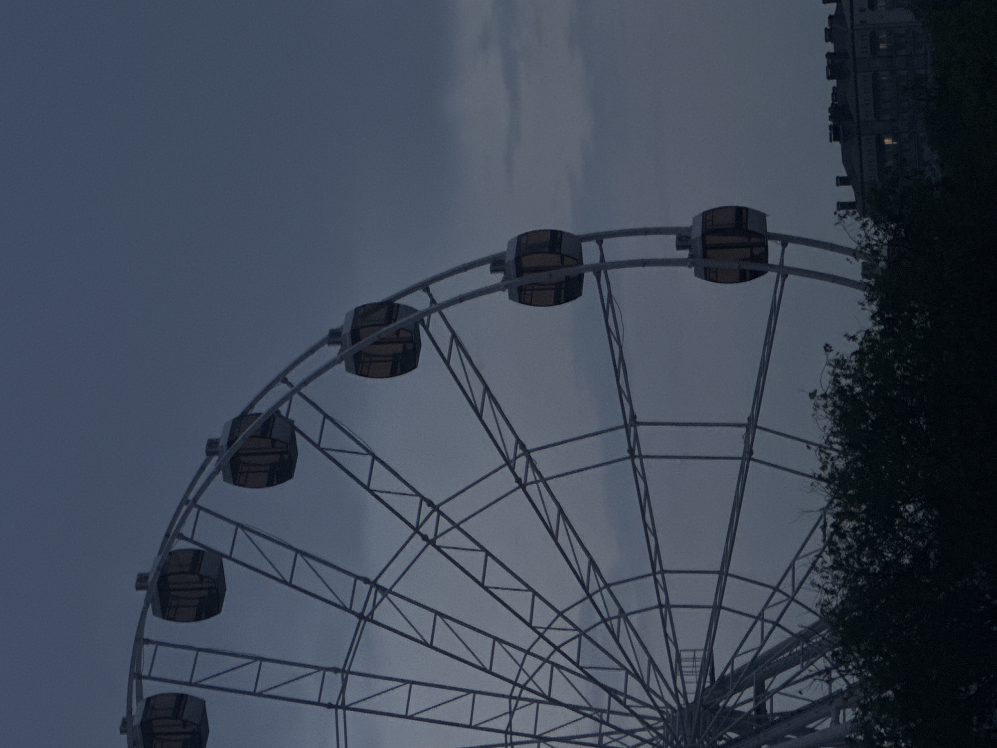 The image shows a portion of a Ferris wheel against a cloudy, dark sky. The wheel's structure is metallic with passenger gondolas attached. Part of a building is visible in the background, to the upper right of the wheel, as well as the top of a tree. This Ferris wheel is located in Yevpatoriya, Ukraine.
