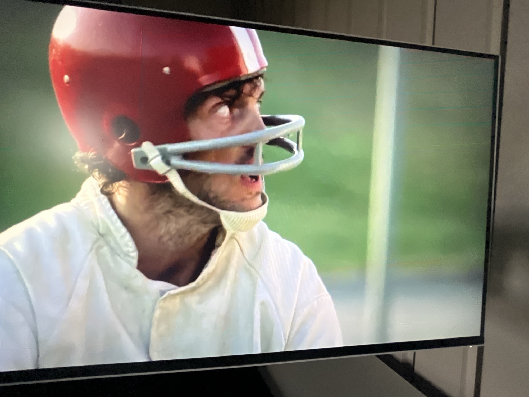 The image shows a close-up of a person wearing a red and white striped football helmet and a white garment, likely a uniform. The person's face is partially visible, and they appear to be looking to the side with their mouth slightly open. The background is a blurred green and white area, suggesting an outdoor setting such as a sports field. There are no discernible details in the image that would indicate its location in Varna, Bulgaria. The image is captured on a television screen, with some reflections and horizontal lines visible on the screen itself, indicating it is a photograph of a screen display.