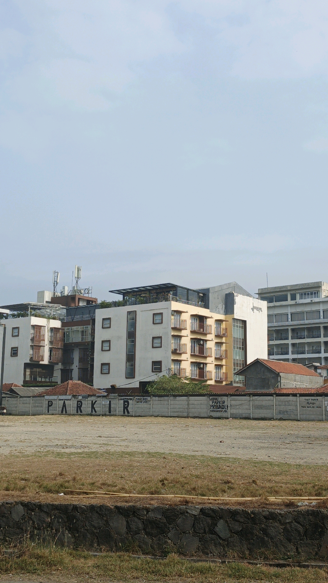 The image shows a view of a street in Desa Pagandaran, Indonesia, with several buildings and a vacant lot in the foreground. In the middle ground, there is a row of buildings, including what appears to be a hotel or apartment complex with multiple floors, balconies, and windows. Some of the buildings have antennas on their roofs. A concrete wall with the word "PARKIR" (Indonesian for parking) stenciled on it runs across the middle of the image. The vacant lot in the foreground is covered with dry grass and dirt, with a stone wall at the very bottom of the frame. The sky is overcast and light blue. There are no people visible in the image.