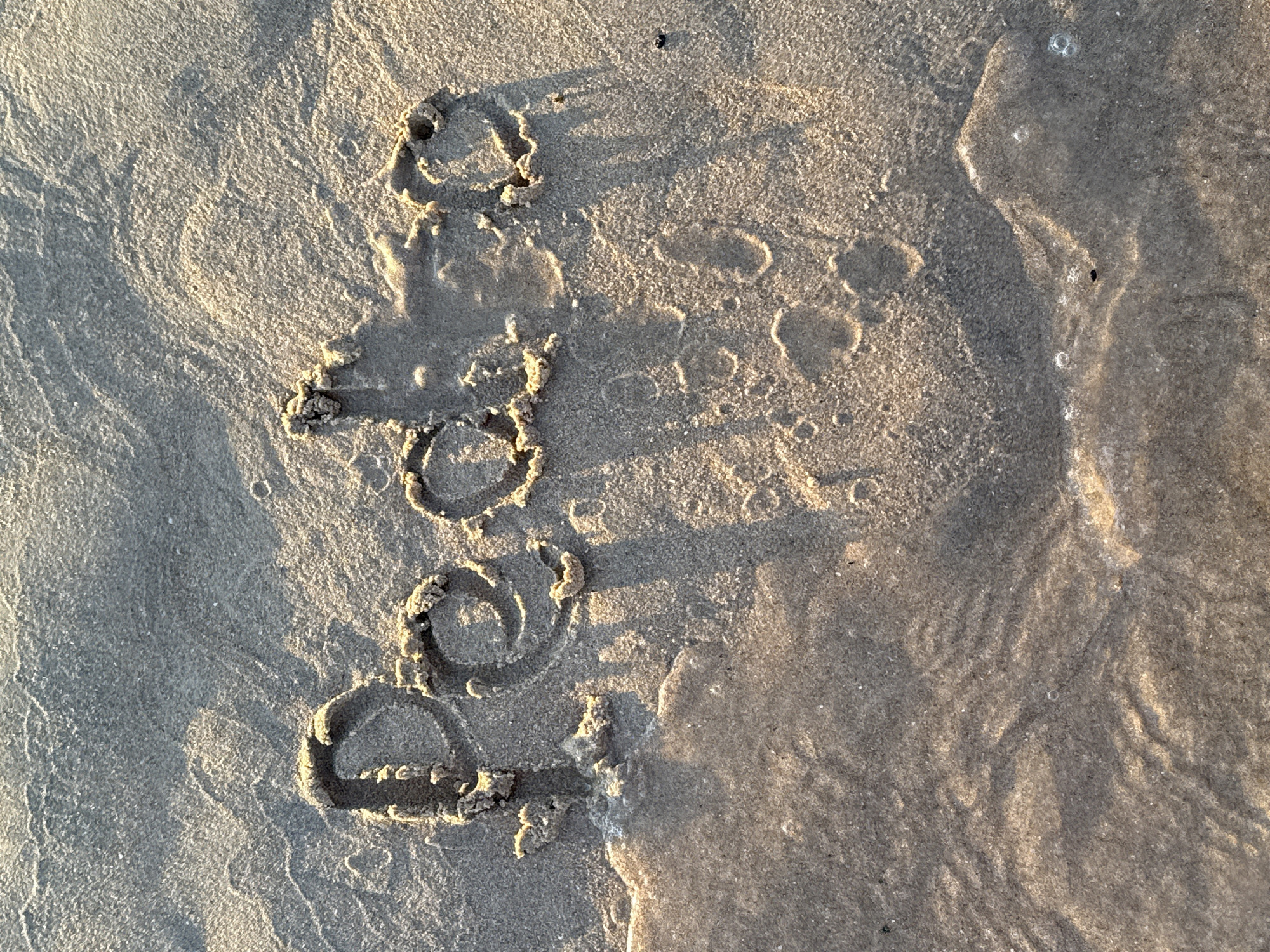 The image shows the word "Peace" written in wet sand on a beach in Cascais, Portugal. The sun casts long shadows from the letters, suggesting it is either early morning or late afternoon. The tide is coming in, with a thin layer of water covering the lower portion of the sand and obscuring some of the lettering. Small bubbles are visible in the water. The sand has a granular texture, and there are some ripples and indentations, indicating natural beach formations or previous activity. The lighting is warm and golden, highlighting the contours of the sand. No people are visible in the image.