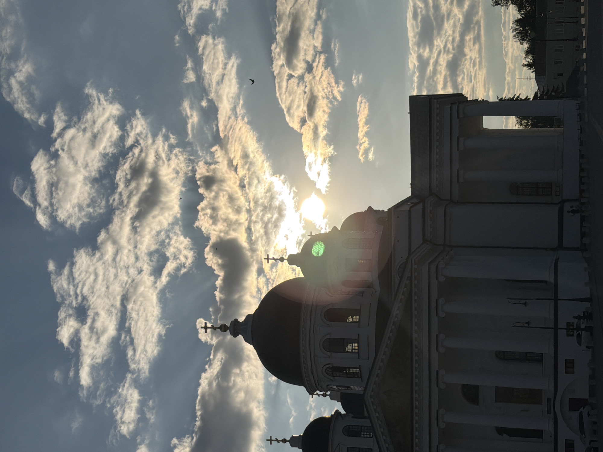 The image shows a view of the Assumption Cathedral in Arzamas, Russia, at sunset. The cathedral's domes and crosses are silhouetted against a dramatic sky filled with clouds illuminated by the setting sun. Rays of light break through the clouds, creating a halo effect around the sun. The facade of the cathedral, with its columns and arched windows, is visible in the foreground. In the sky, two birds are captured in flight. The surrounding environment includes some trees and buildings in the distance, as well as a few cars and streetlights at the bottom of the frame. The overall scene conveys a sense of grandeur and atmospheric beauty.