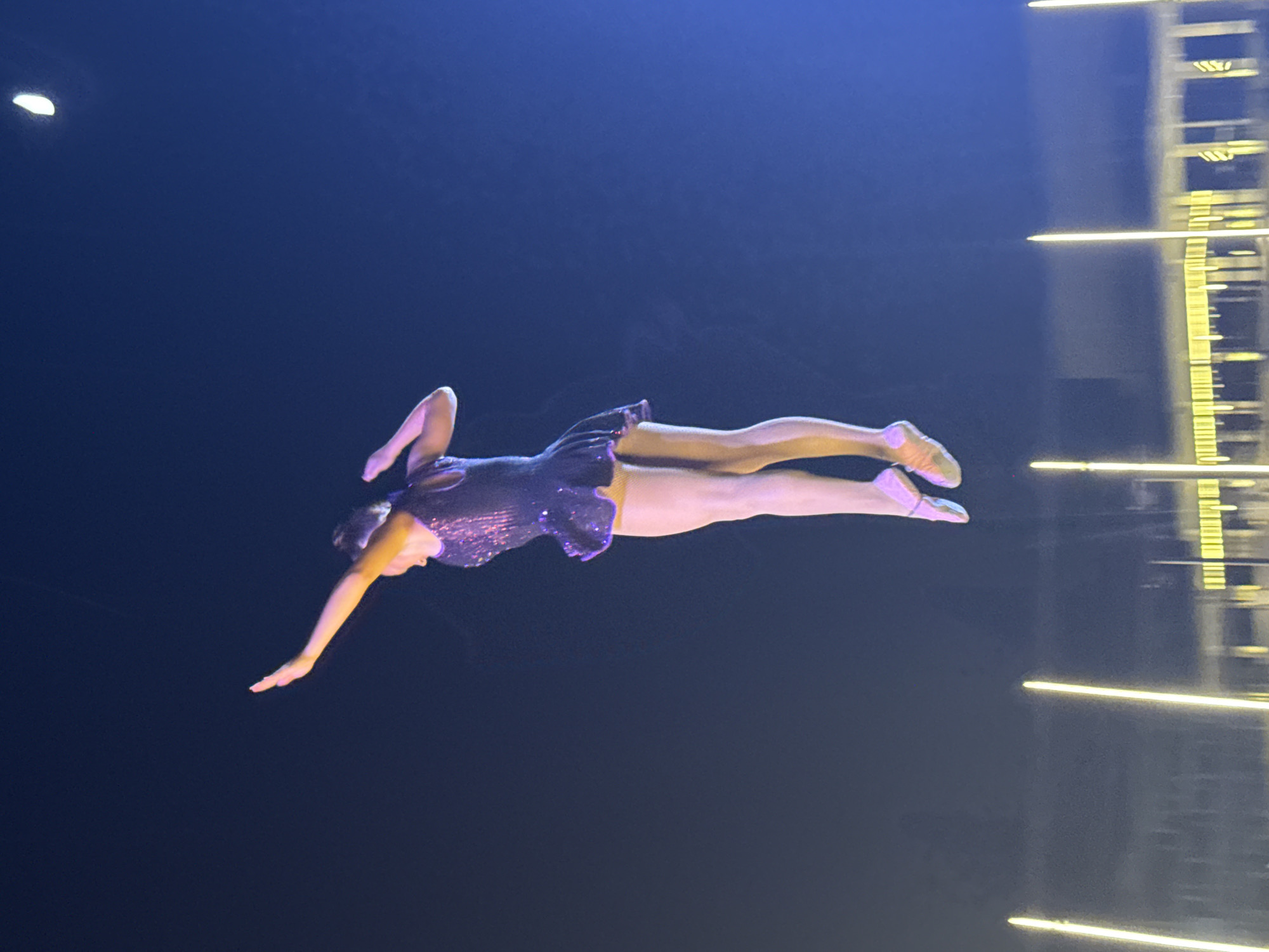 An ice skater in a sequined purple dress and pink skates is captured mid-air during a jump against a dark blue night sky. Her arms are extended, one upward and outward, the other bent at the elbow. A sliver of the moon is visible in the upper right corner. Below the skater, out of focus, are lights that appear to be part of a stage or performance area, with tall, thin vertical lights and a horizontal structure with multiple lights. The background is dark, suggesting an outdoor nighttime event in Sharm El Sheikh, Egypt.