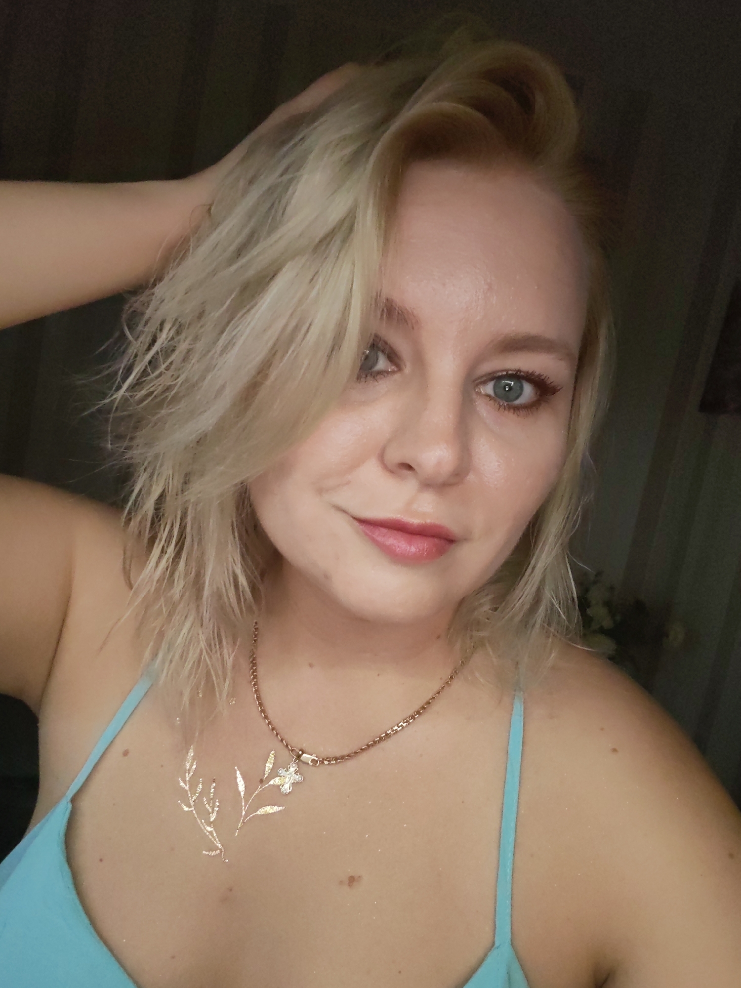 A medium shot captures a Caucasian woman with blonde, wet hair pulled back with her left hand. She is looking directly at the camera, her head tilted slightly. Her eyes are light blue, and she wears subtle makeup, including eyeliner and pink lipstick. A delicate rose gold chain necklace with a flower charm rests on her chest, above a gold temporary tattoo of a branch with a flower. She is wearing a light blue camisole top. The background is dark and out of focus, with vertical stripes visible on the right, suggesting an indoor setting, possibly a bedroom or bathroom. There are no visible indicators of the specific location in Izhevsk, Russia, beyond the general context of an indoor environment.